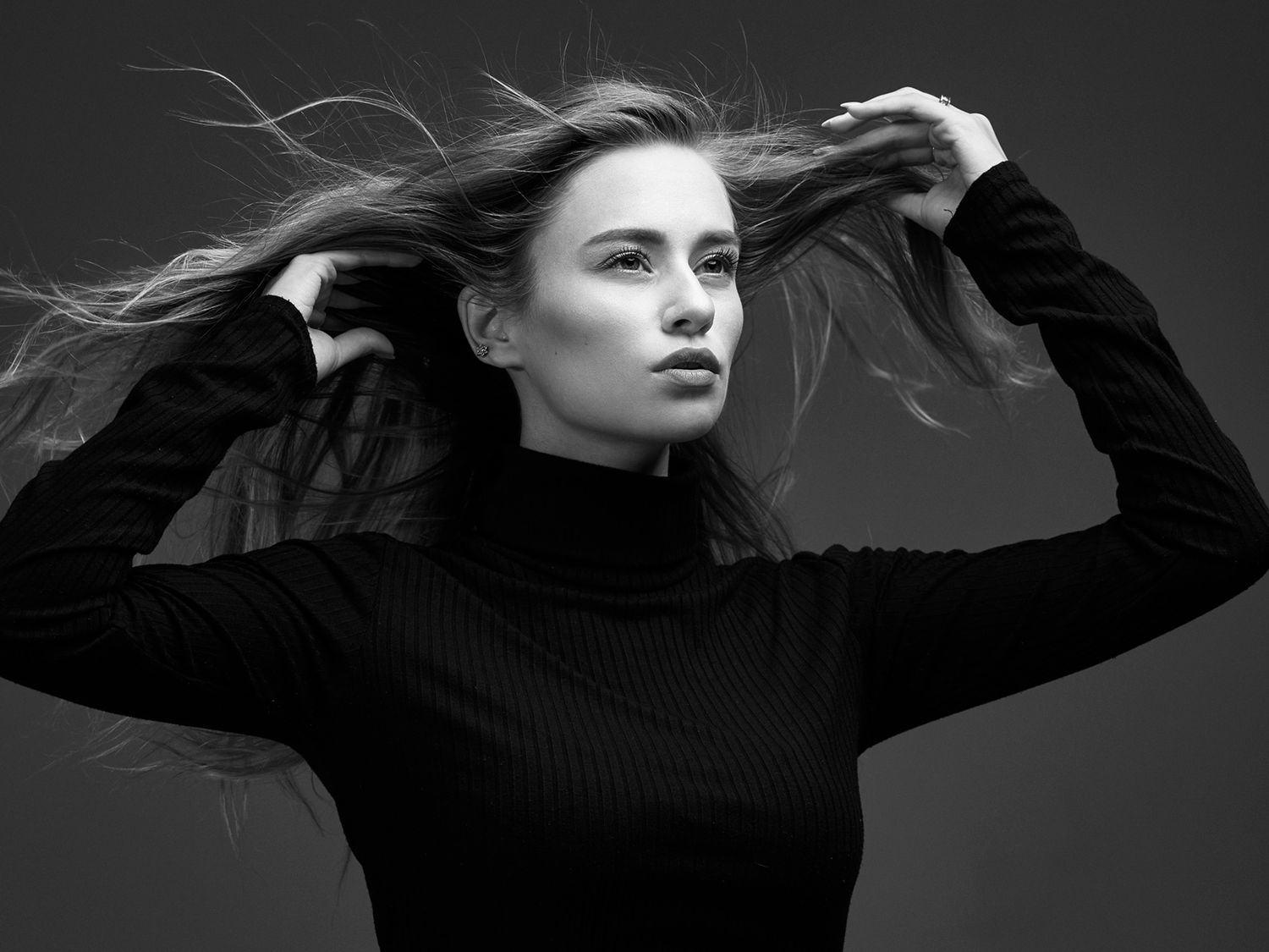 Dramatic black and white portrait with flowing hair and black turtleneck against dark background.