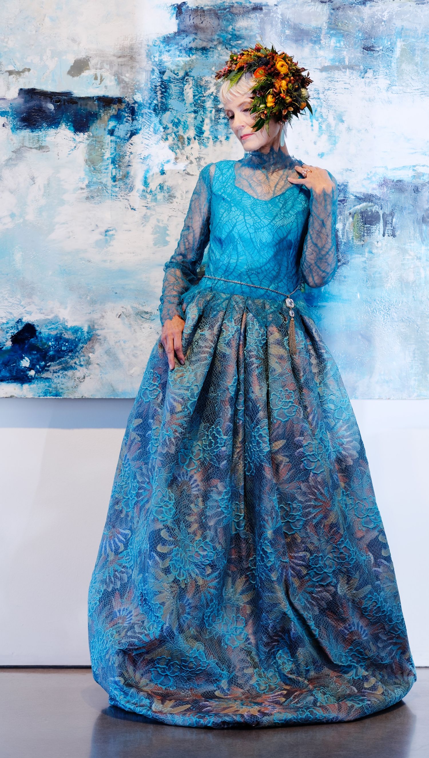 An elegant blue brocade ball gown with floral patterns is showcased against an abstract blue and white painted backdrop.