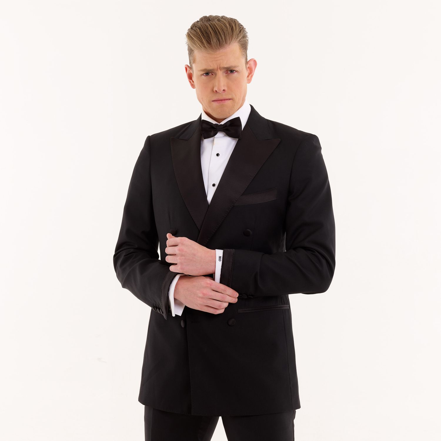 Person in black tuxedo adjusts bow tie against white background in elegant formal pose.