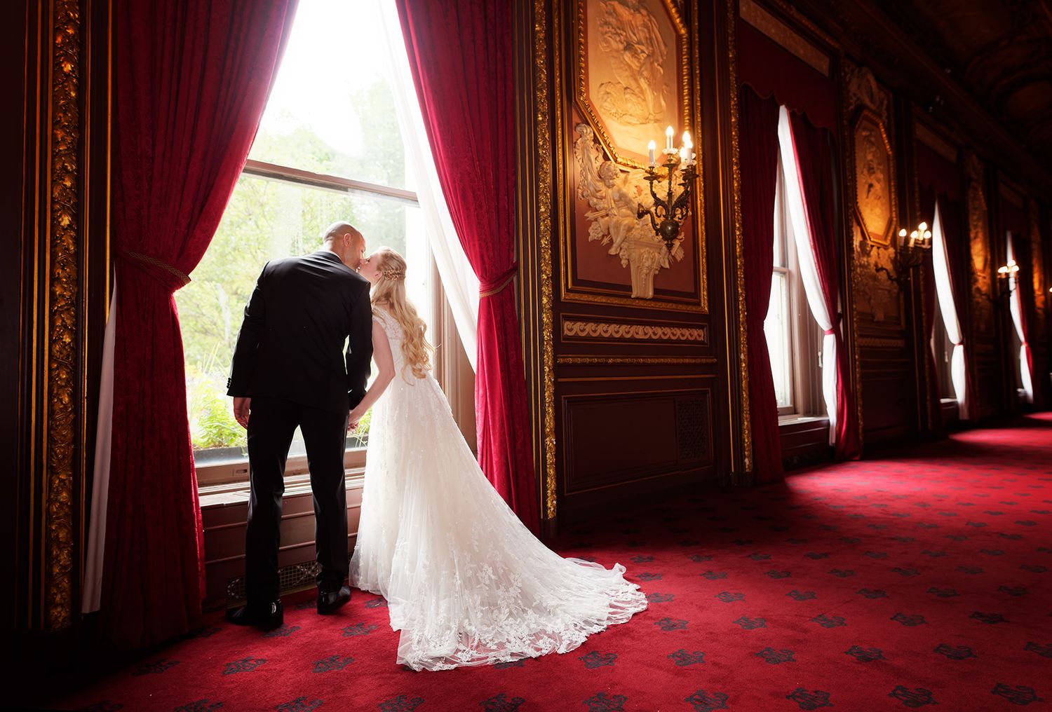 A couple looks out tall windows in an opulent vintage mansion with red carpet and ornate gold walls.