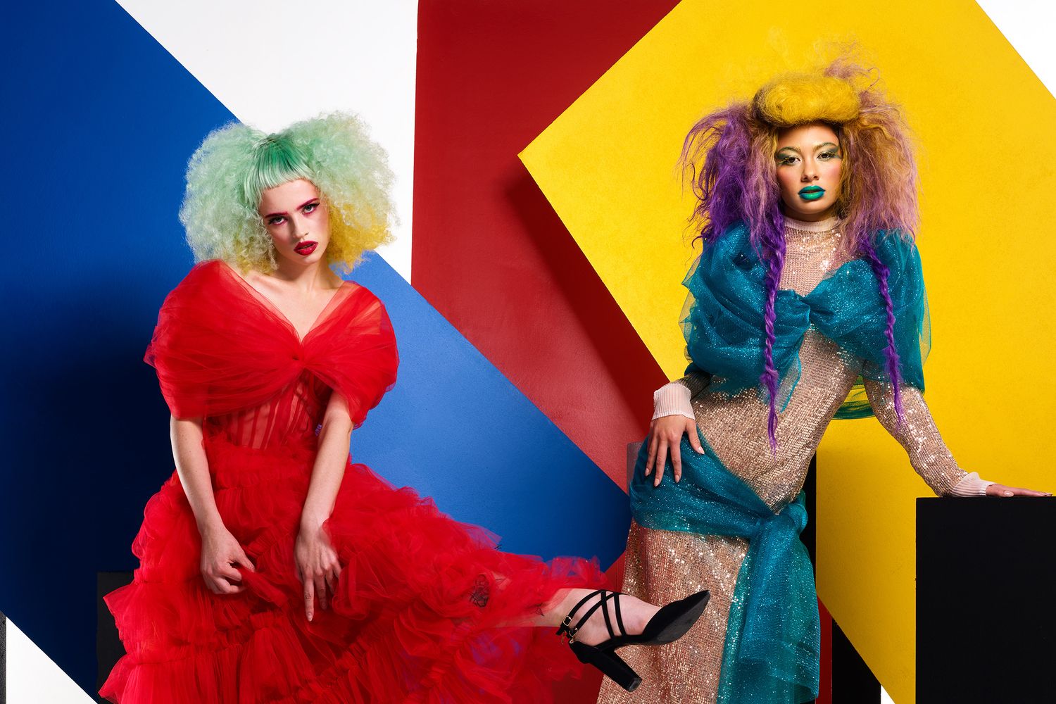 Two models in colorful outfits pose in front of geometric color block backdrop, one in red and one in teal.