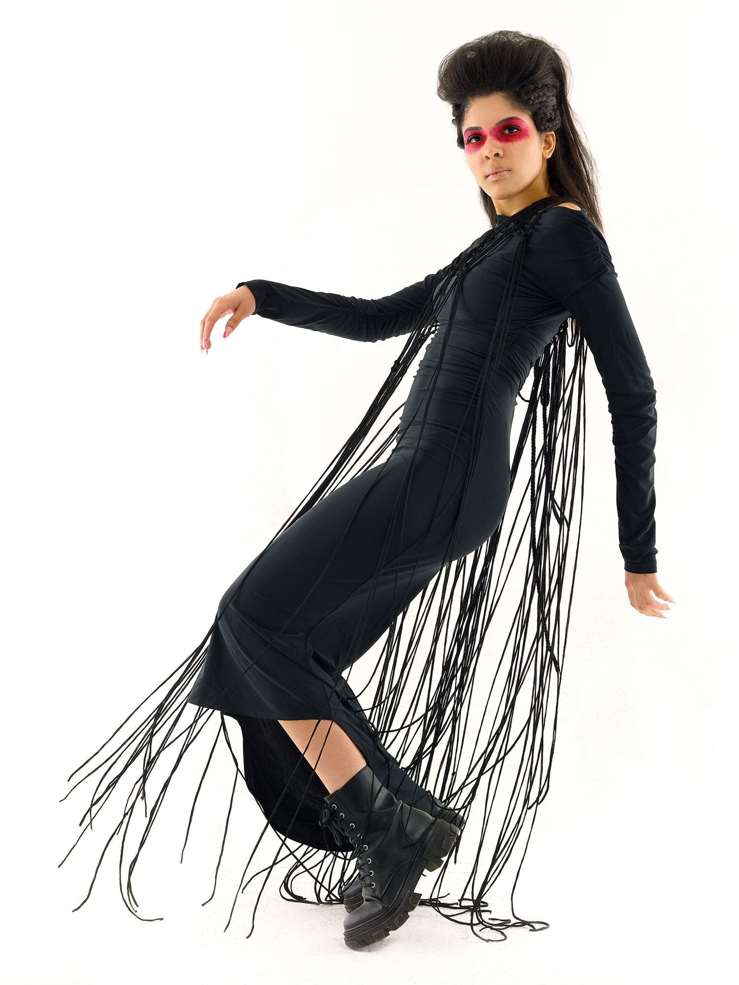 Figure in dramatic black fringe dress and combat boots strikes a pose against white background wearing red sunglasses.