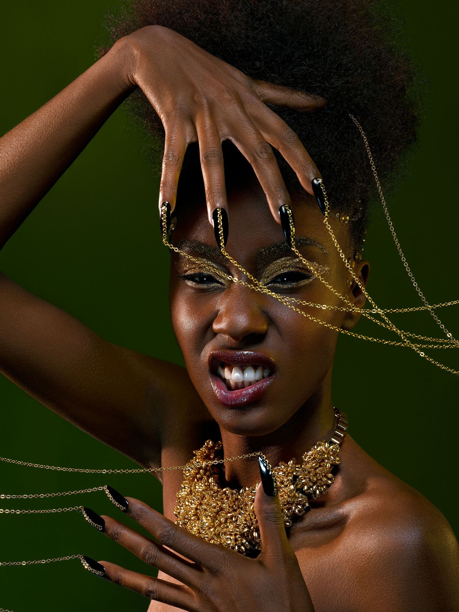 Creative fashion portrait with gold glitter makeup, statement necklace and dramatic hand pose against dark green background.