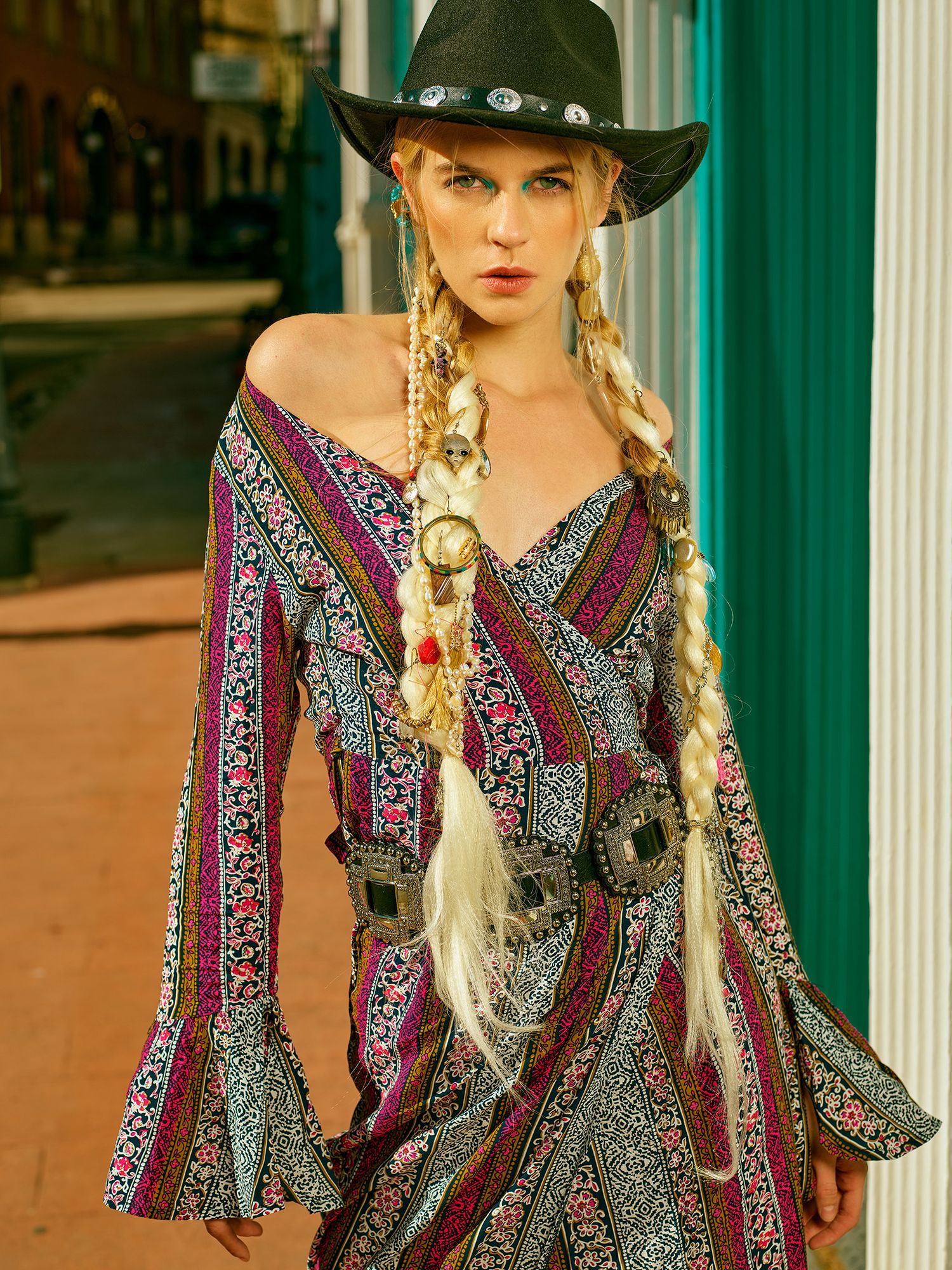A fashion model in cowboy hat wearing colorful boho dress with bell sleeves against turquoise building columns.