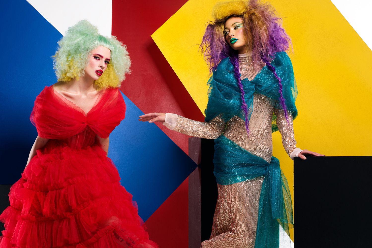 Two models in dramatic costumes pose against colorful geometric backdrop with vibrant wigs.