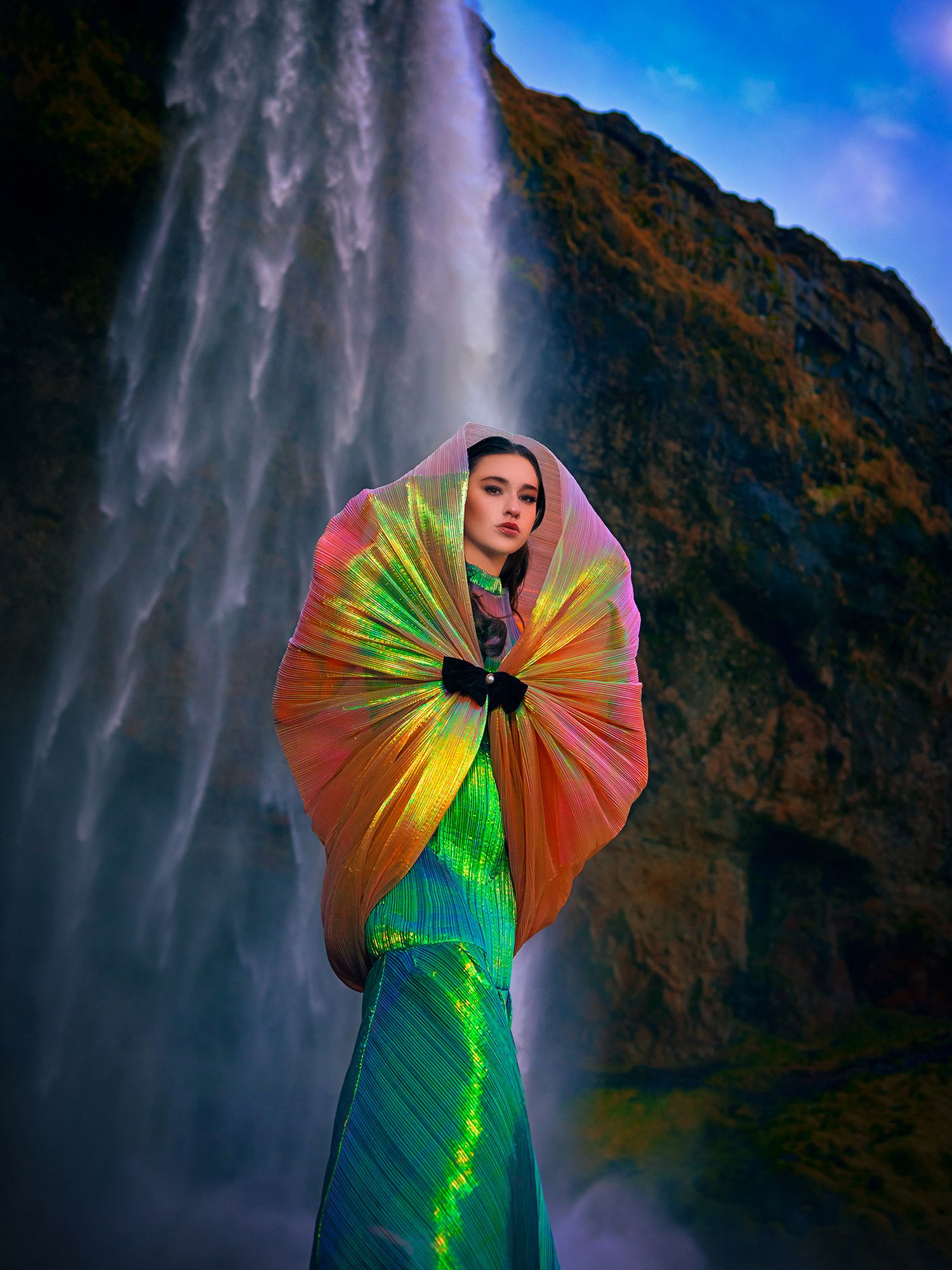 Figure in a vibrant rainbow dress poses dramatically against a towering waterfall backdrop in scenic Iceland.