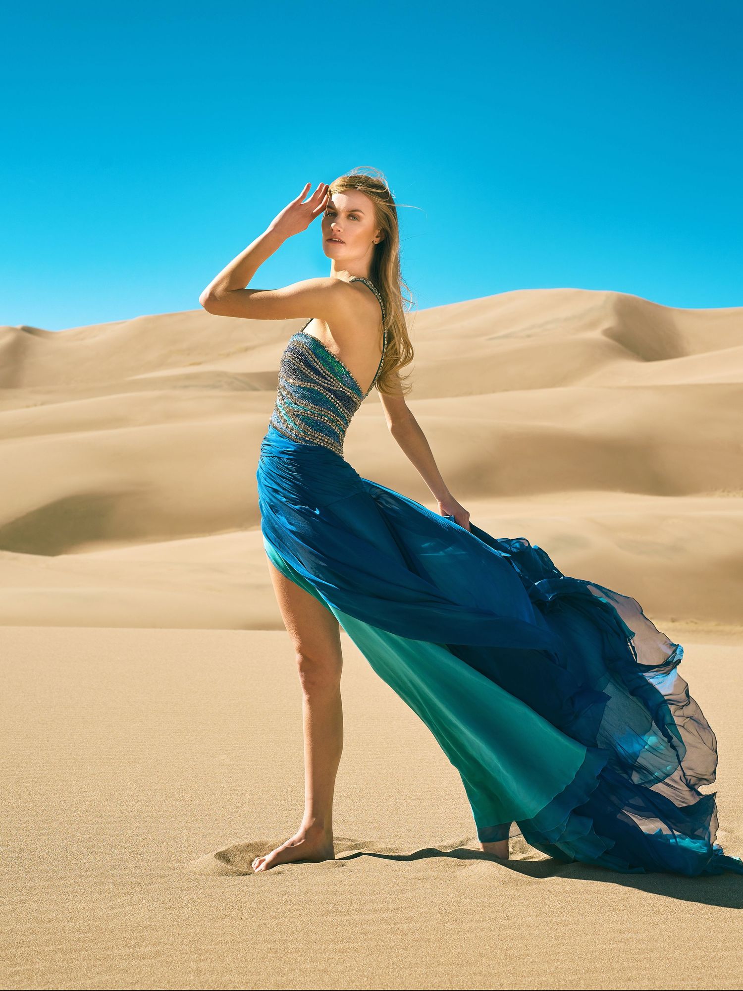 A person in a flowing blue turquoise gown stands barefoot in sand dunes against bright blue sky in desert photoshoot.