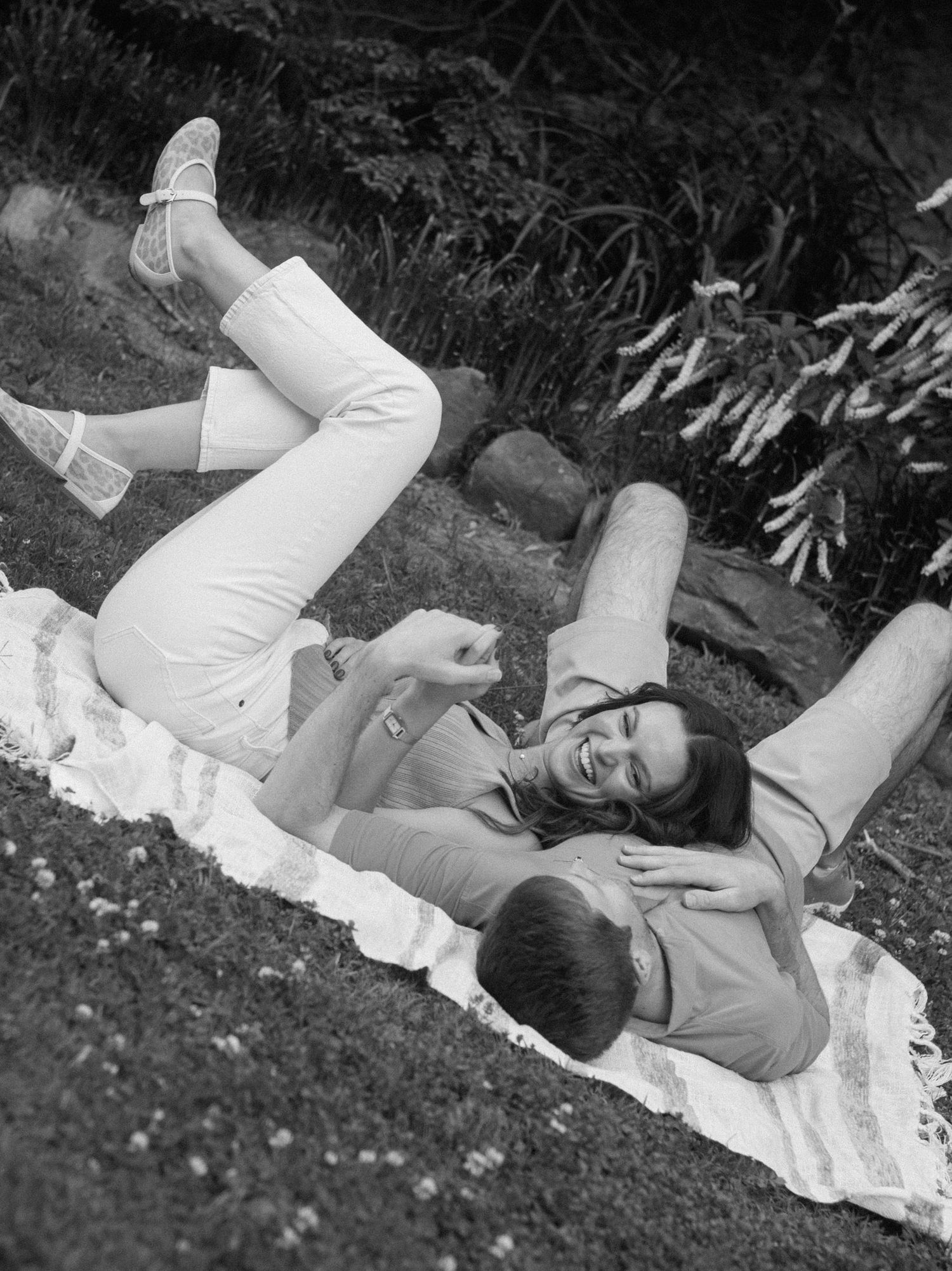 A vintage black and white photo shows two people relaxing on a blanket on the grass, laughing and laying down together.