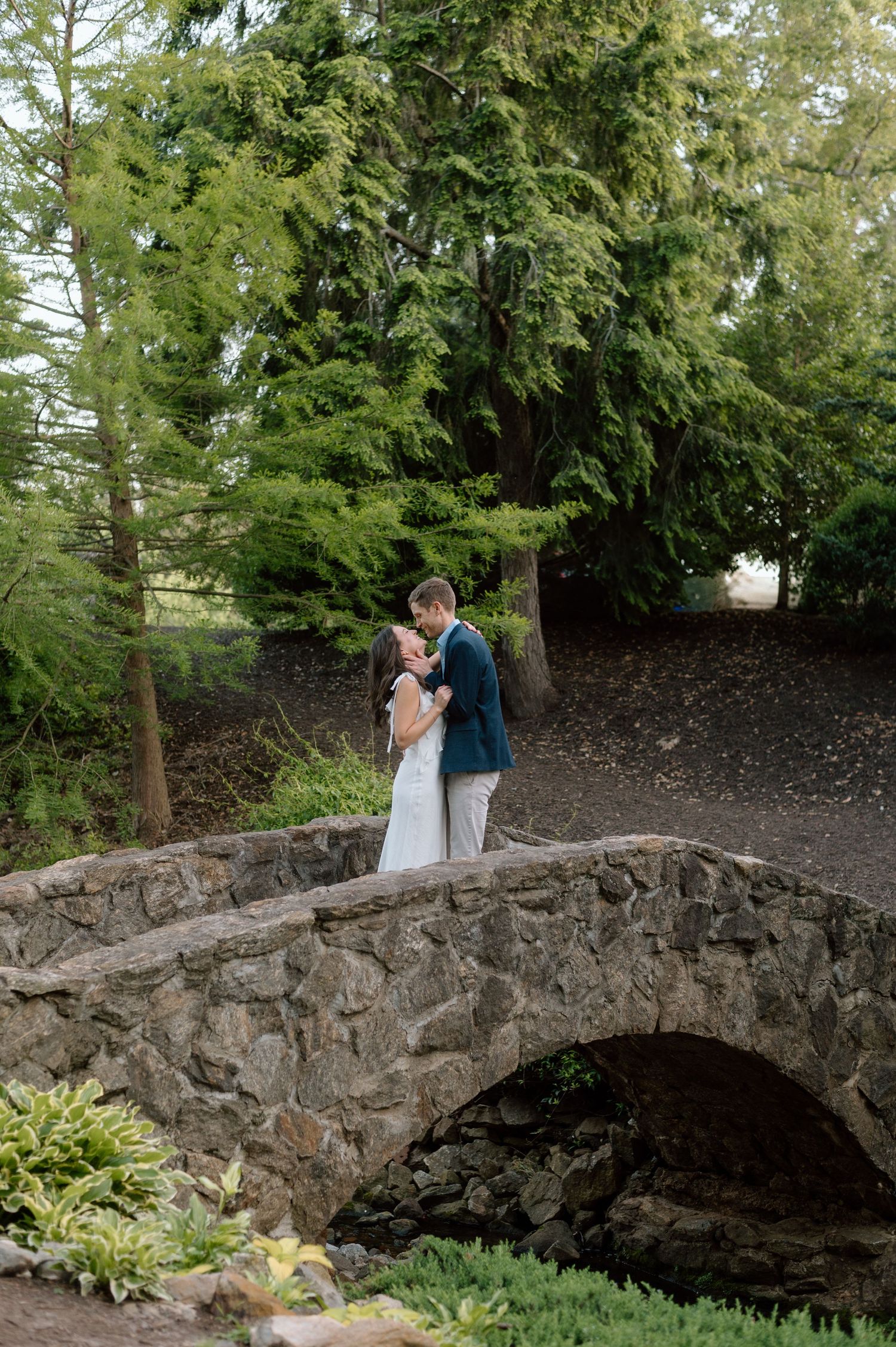 A romantic moment shared between a couple on a rustic stone bridge surrounded by lush evergreen trees.
