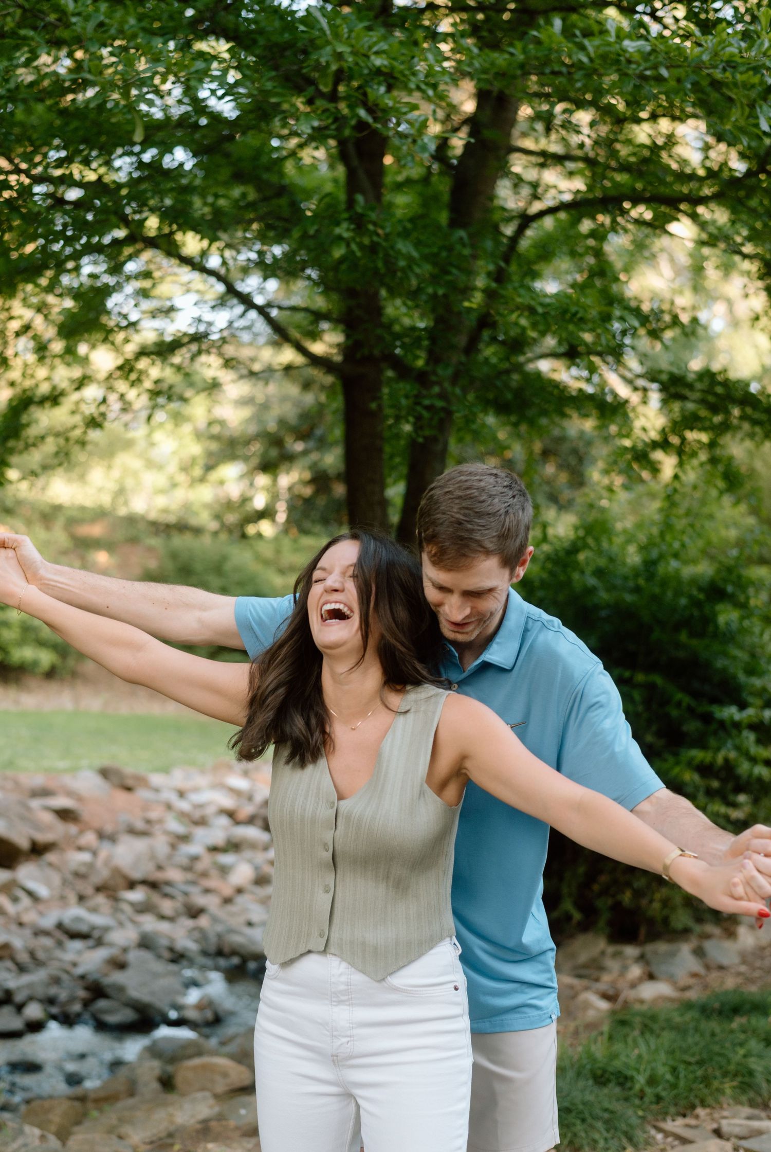 A joyful couple enjoying a playful moment together outdoors near a creek with green trees in the background.