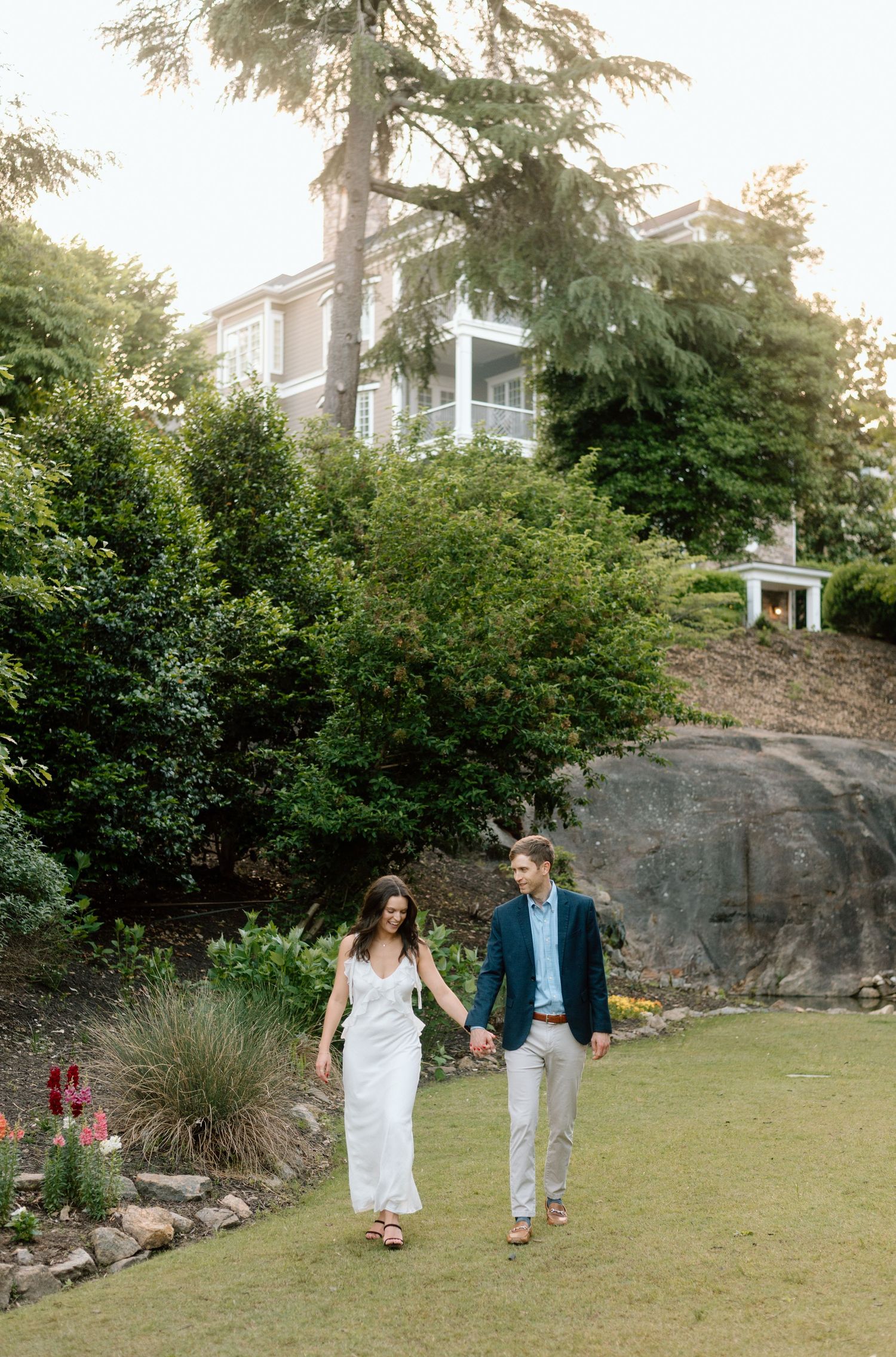 Couple walking hand in hand on grassy path next to rock outcropping with large house on hillside behind them.