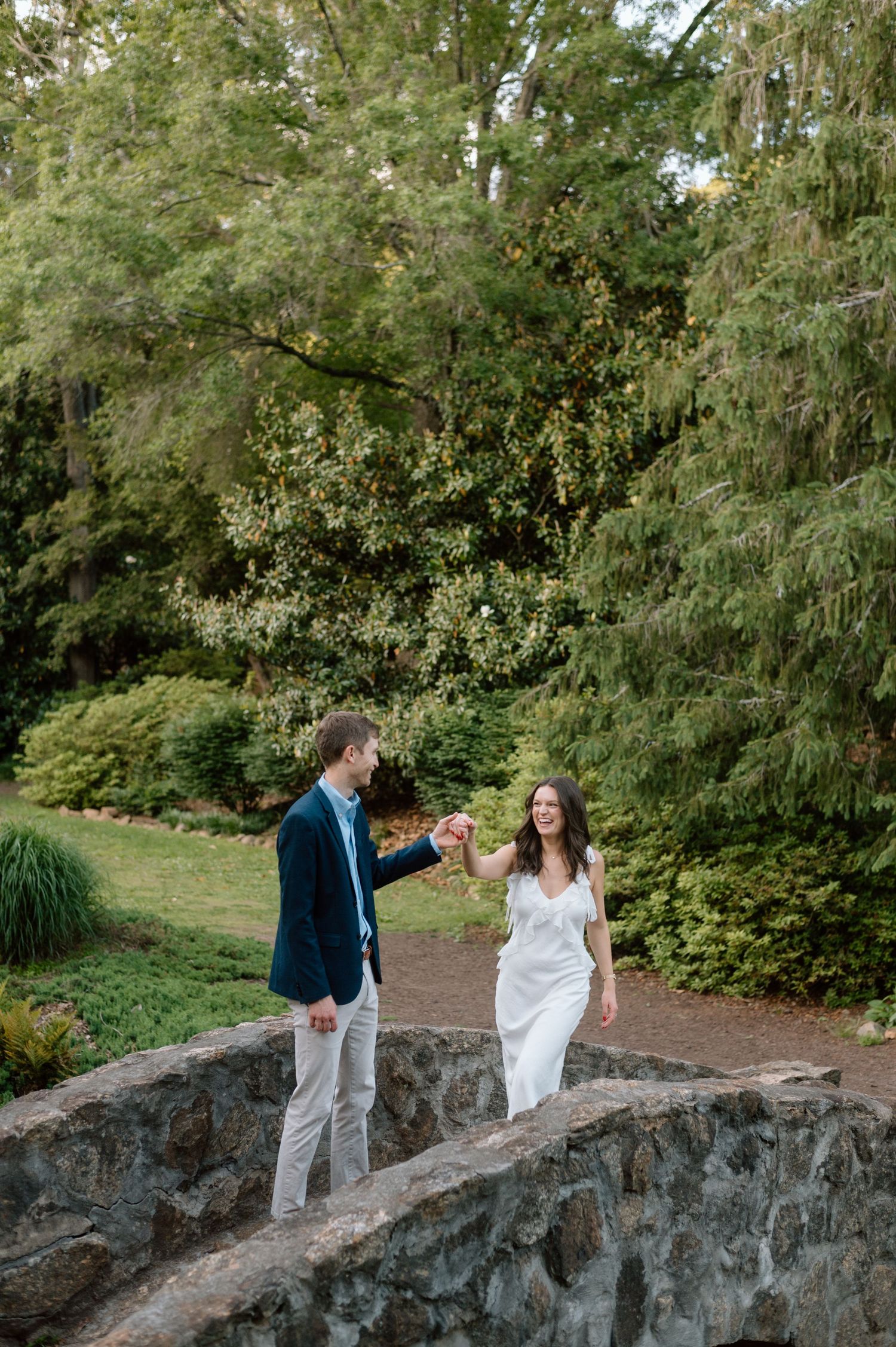 A couple walks together on a stone bridge path surrounded by lush green trees and vegetation in a garden setting.