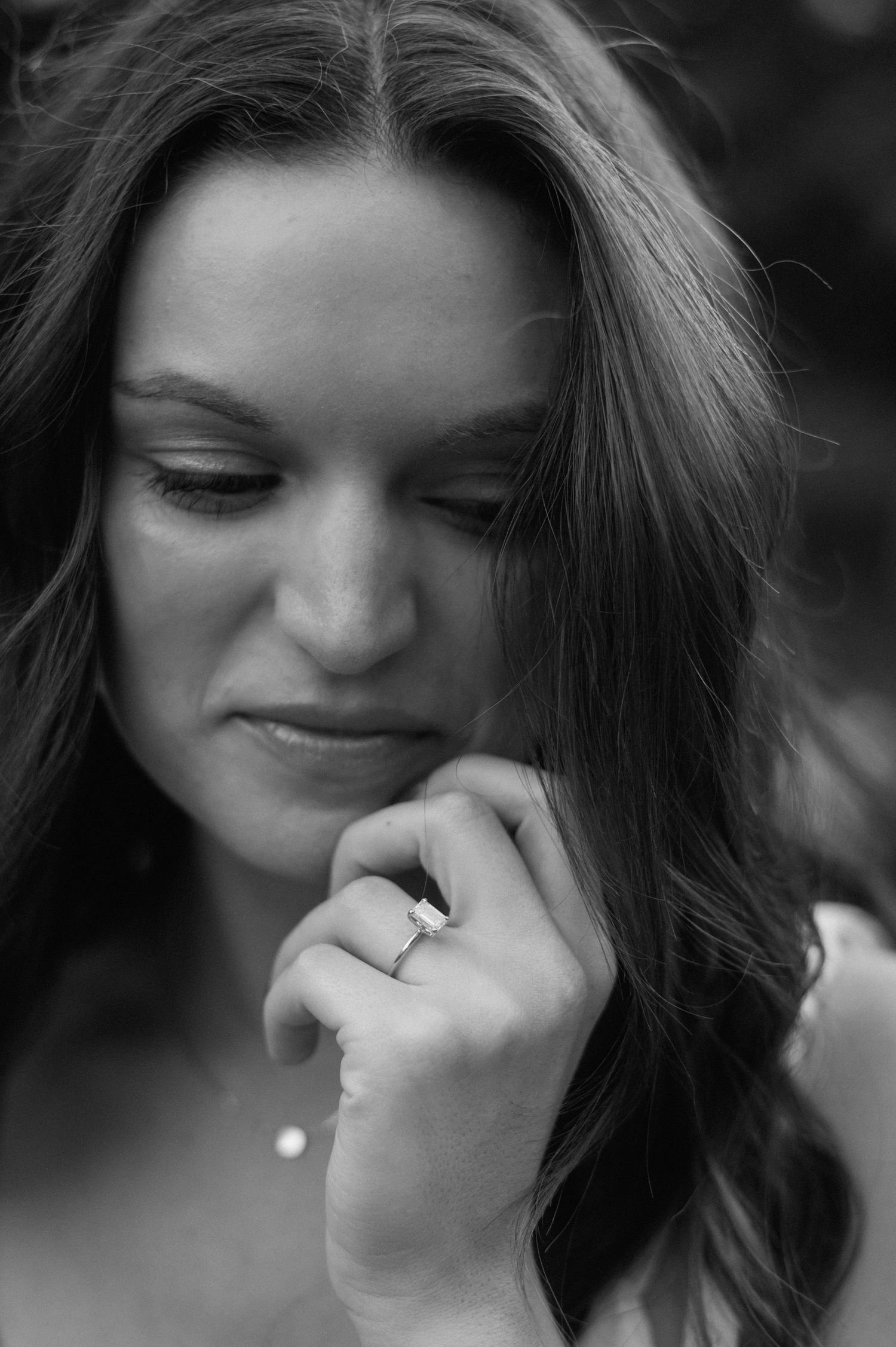 Black and white portrait showing a downward glance and gentle smile while displaying an engagement ring.