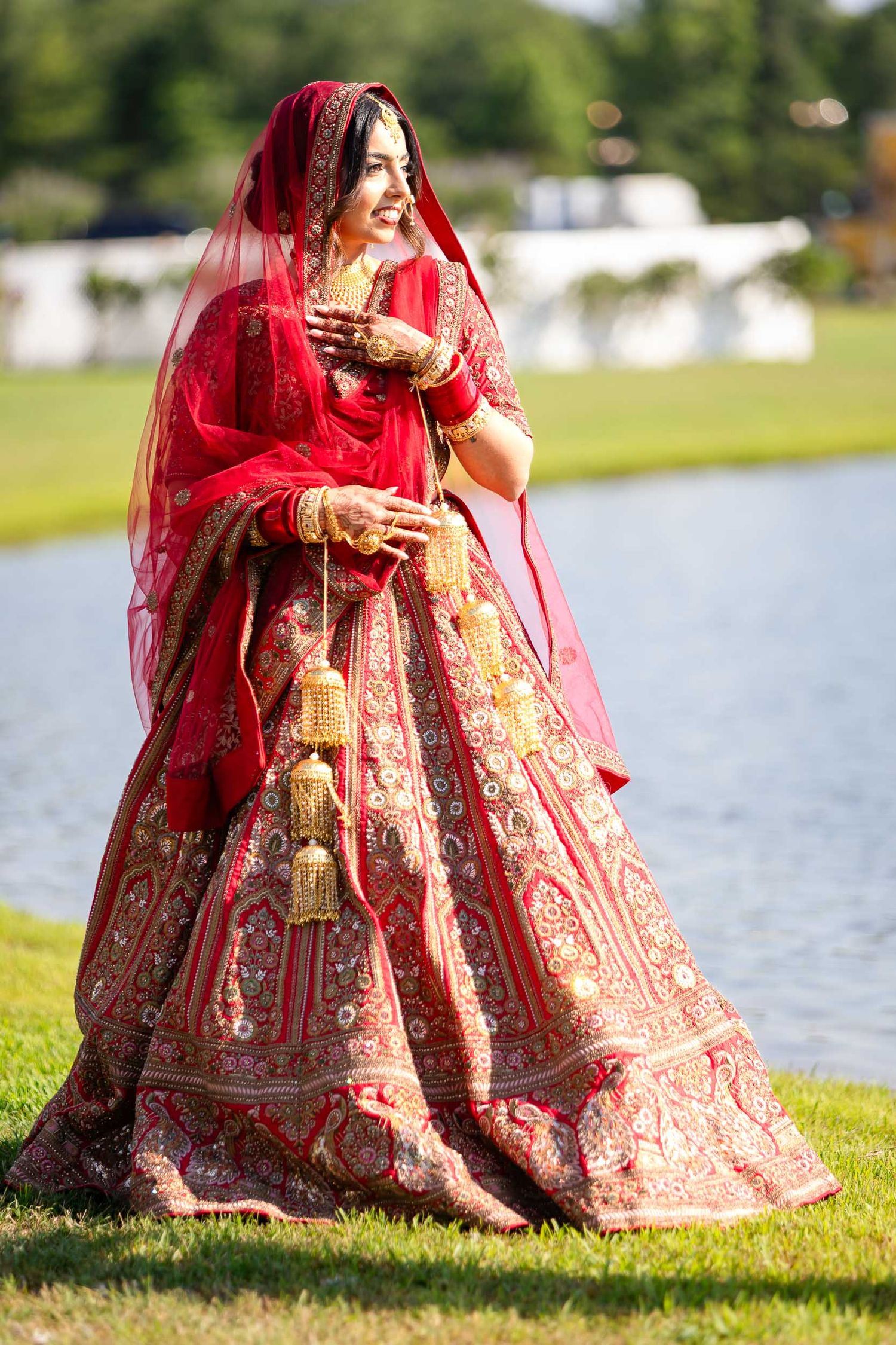 Traditional Indian bride wearing a red and gold embroidered lehenga poses gracefully by a lake on a sunny day.