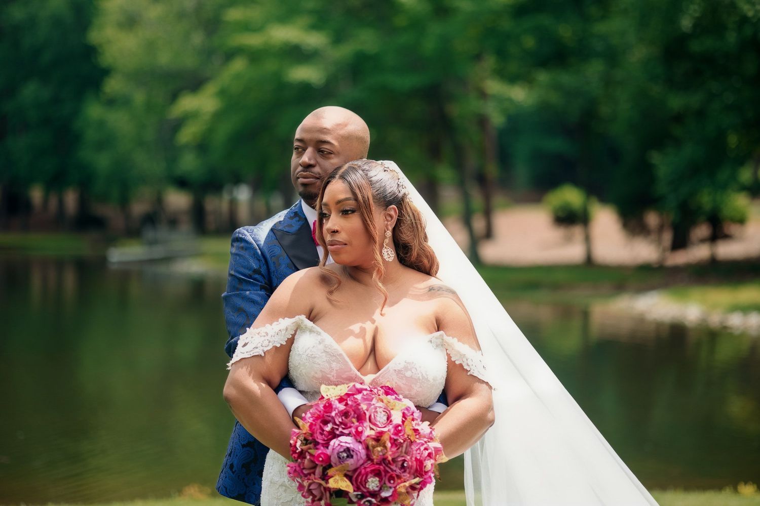 Happy newlywed couple embracing by lake with pink floral bouquet in romantic outdoor setting.