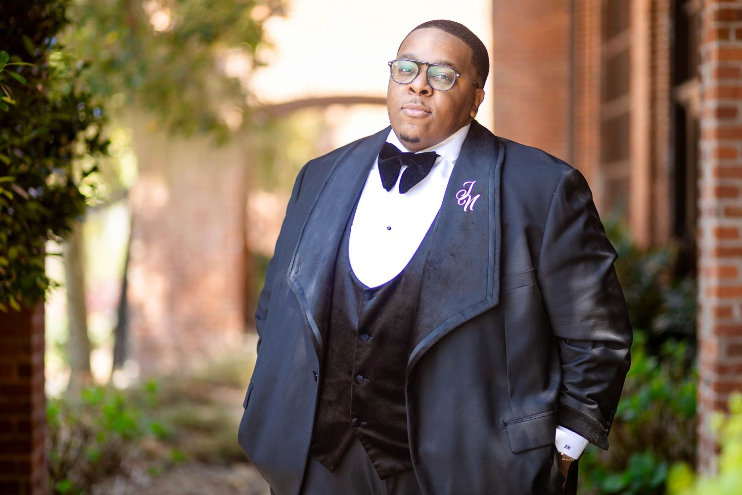 Person in formal black tuxedo with tails and bow tie poses outdoors near brick building.