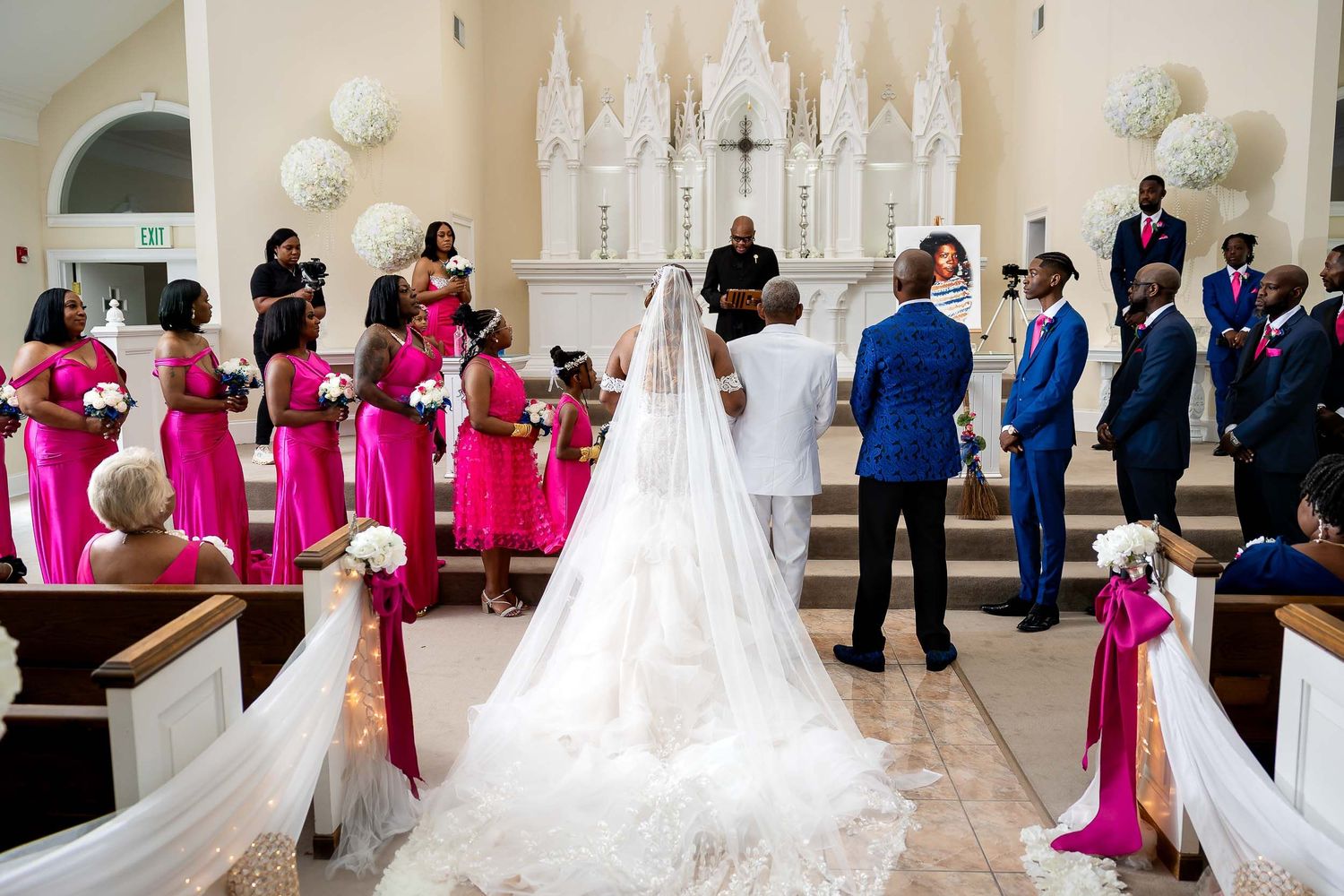 A wedding ceremony in a white church with bridesmaids in bright pink dresses and groomsmen in navy suits gathered at the altar.