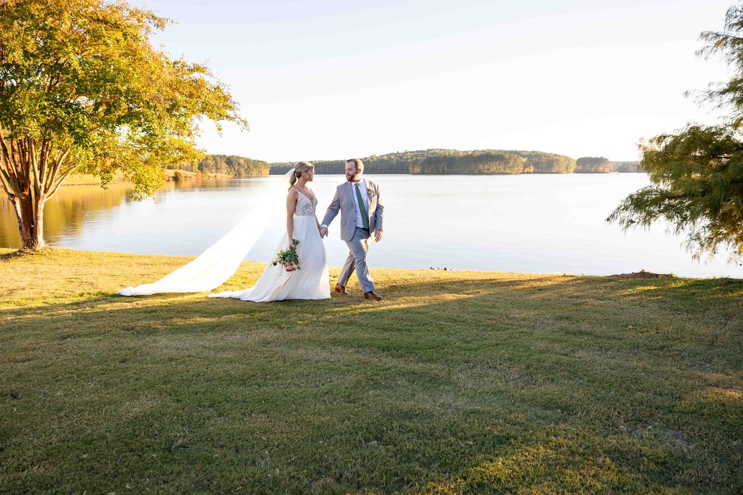 A couple walks together on a grassy lawn overlooking a tranquil lake at sunset during their wedding celebration.