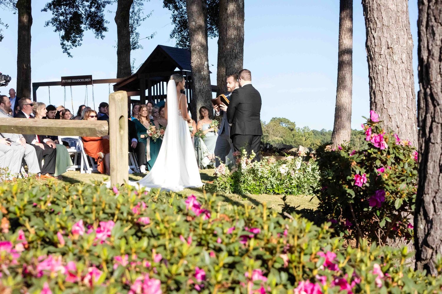 An outdoor wedding ceremony overlooking a lake with pink flowers in the foreground and tall pine trees framing the scene.