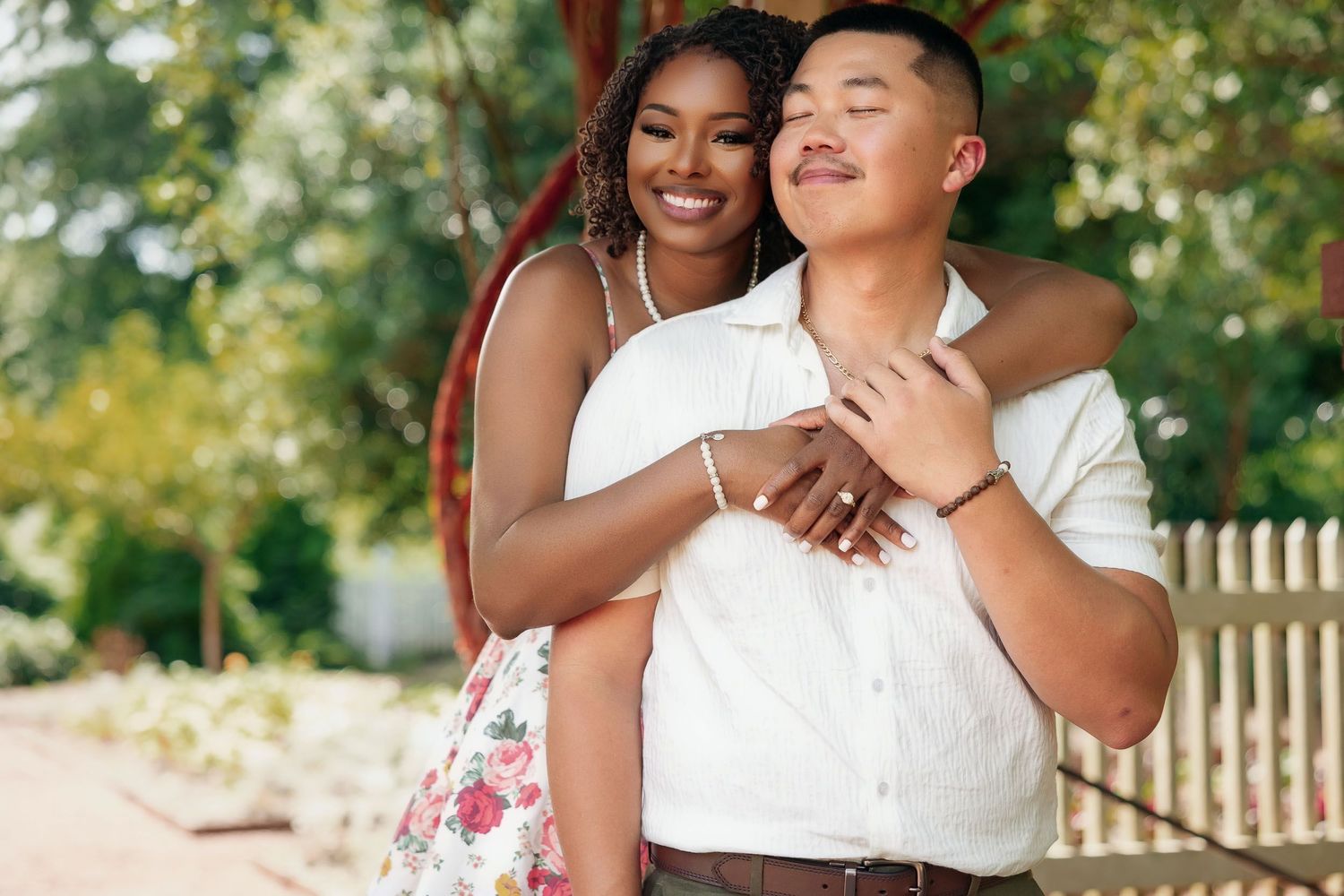 A loving couple embraces while laughing together outdoors on a sunny day with flowers and a fence in the background.