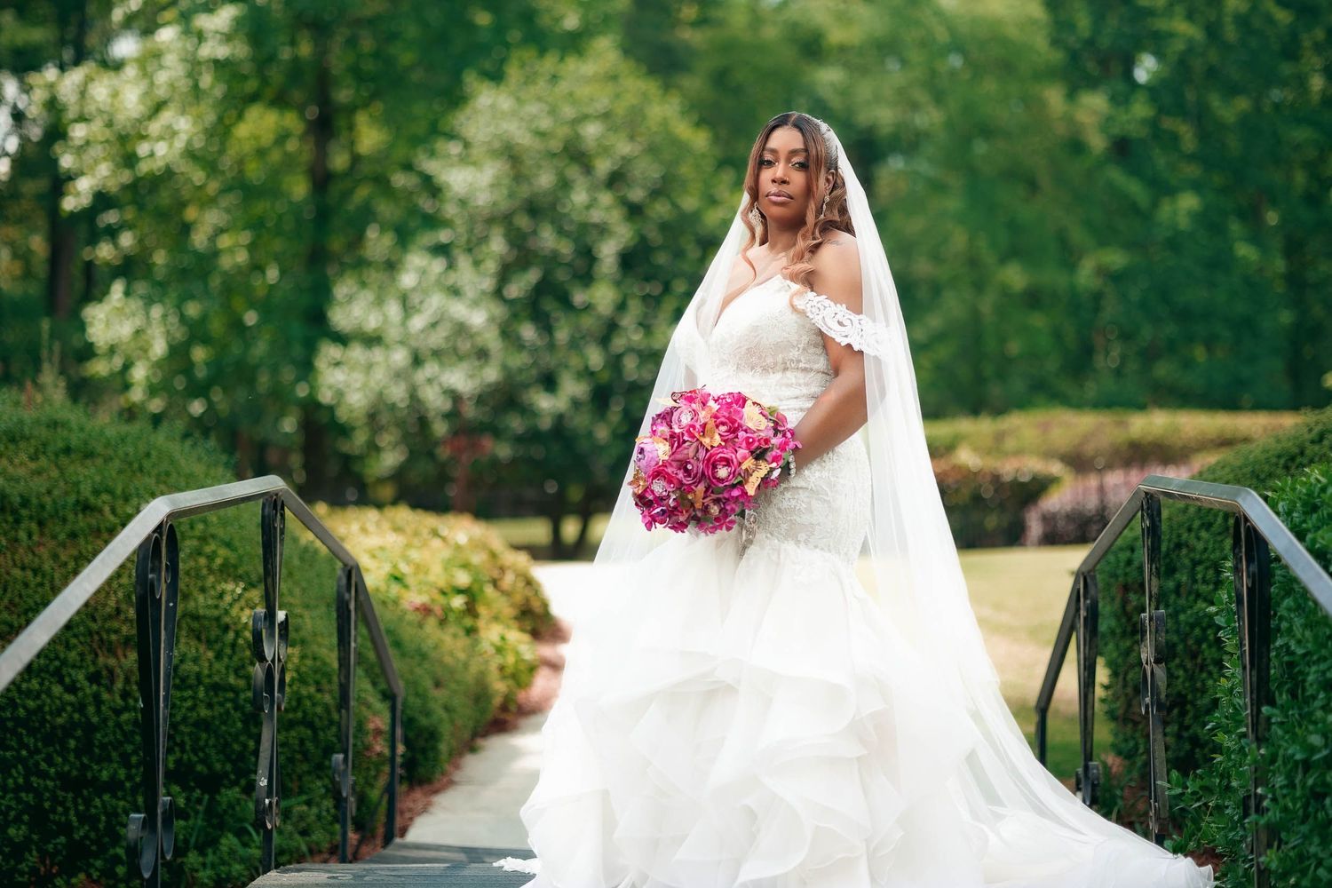 Bride in flowing white gown holds vibrant pink bouquet while standing on garden bridge surrounded by lush greenery.