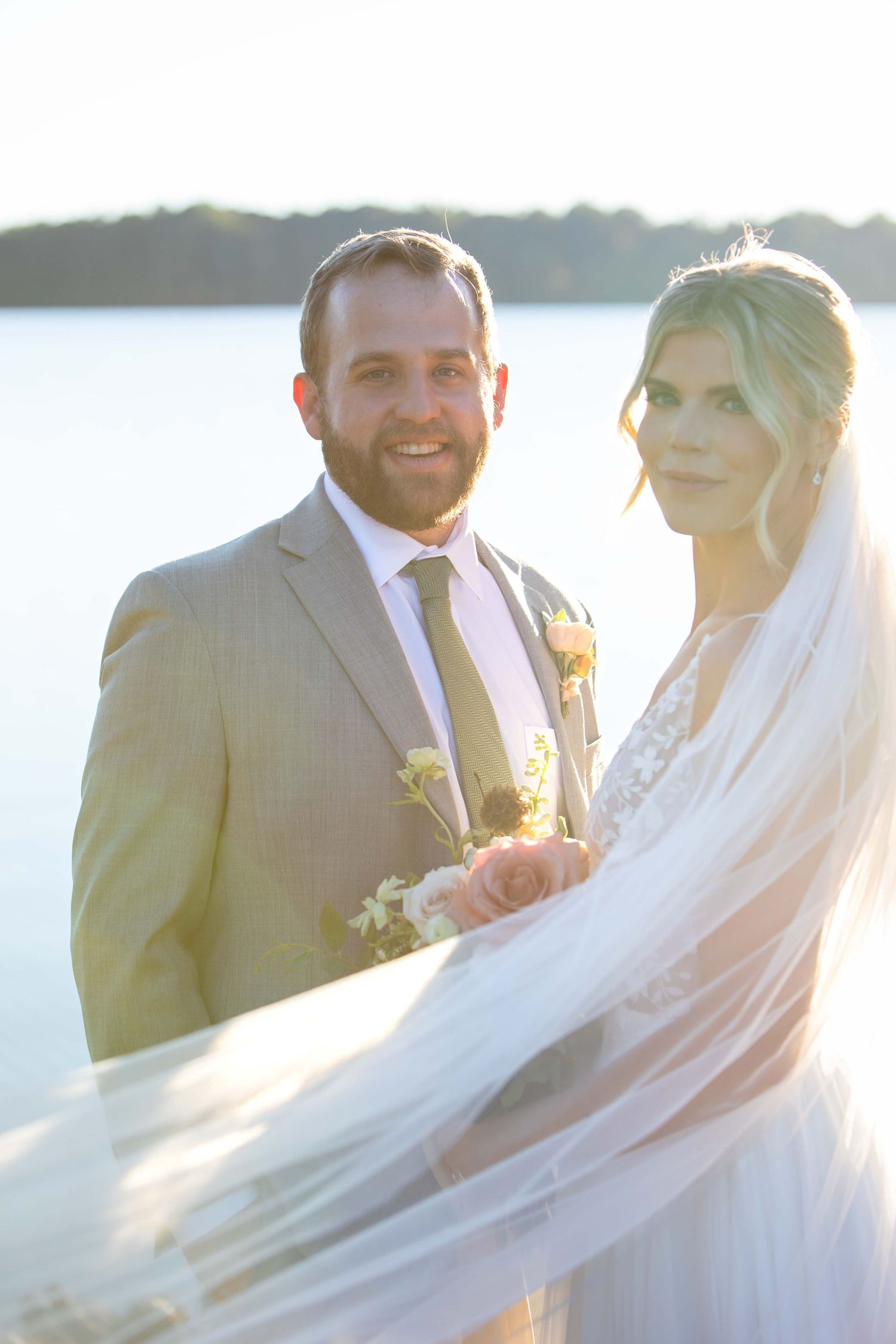 A couple stands together on their wedding day by a lake as a flowing white veil catches the sunlit breeze.