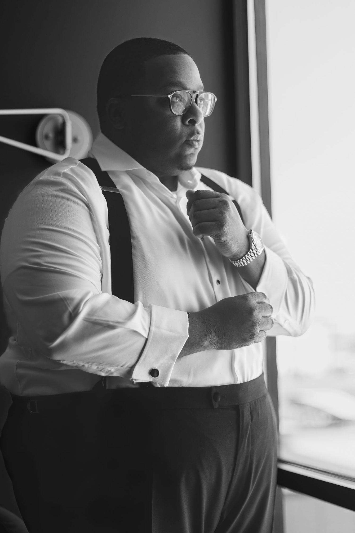 A person wearing glasses and formal attire adjusts their tie while standing by a window in black and white photography.