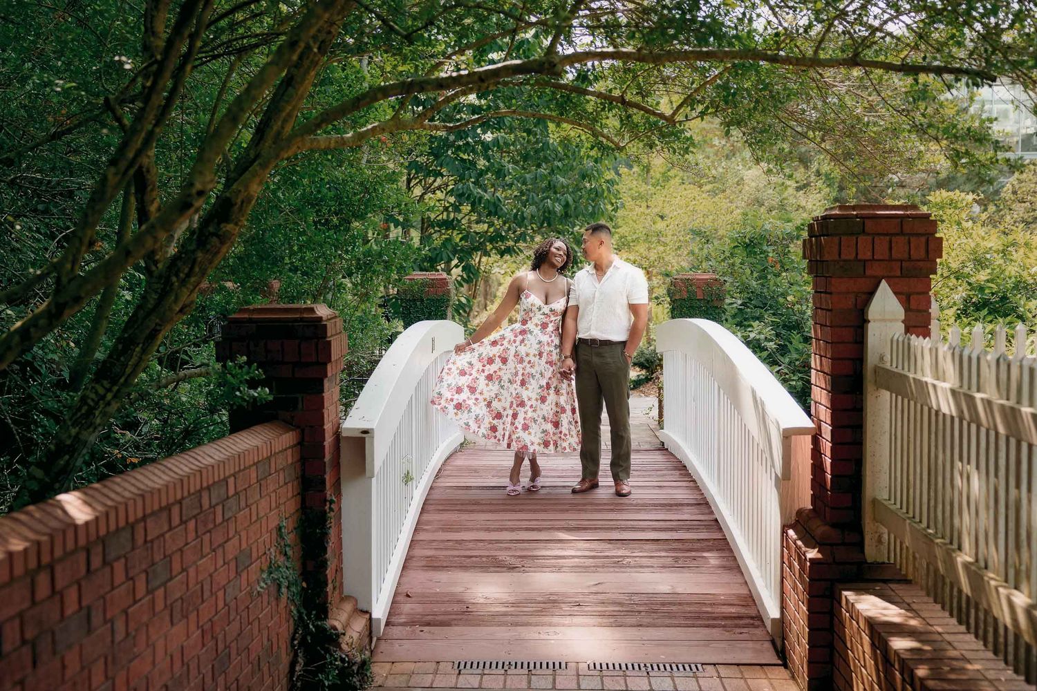 A couple stands together on a white wooden footbridge surrounded by lush green trees and red brick pillars.