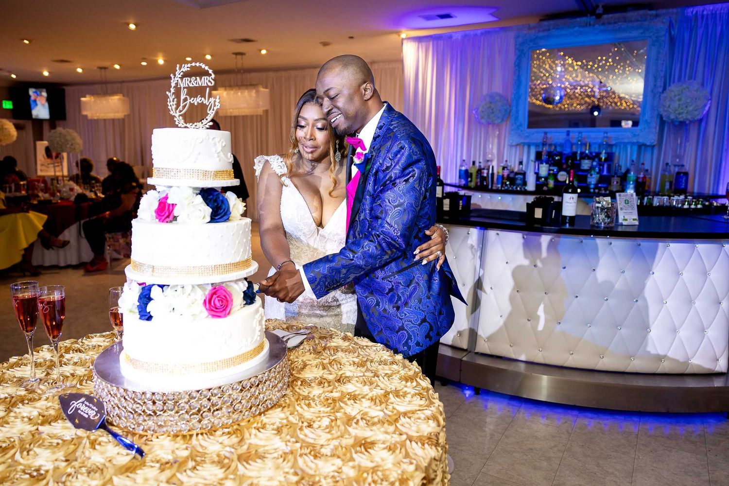 A couple shares a tender moment while cutting their elegant white wedding cake decorated with blue and pink flowers.