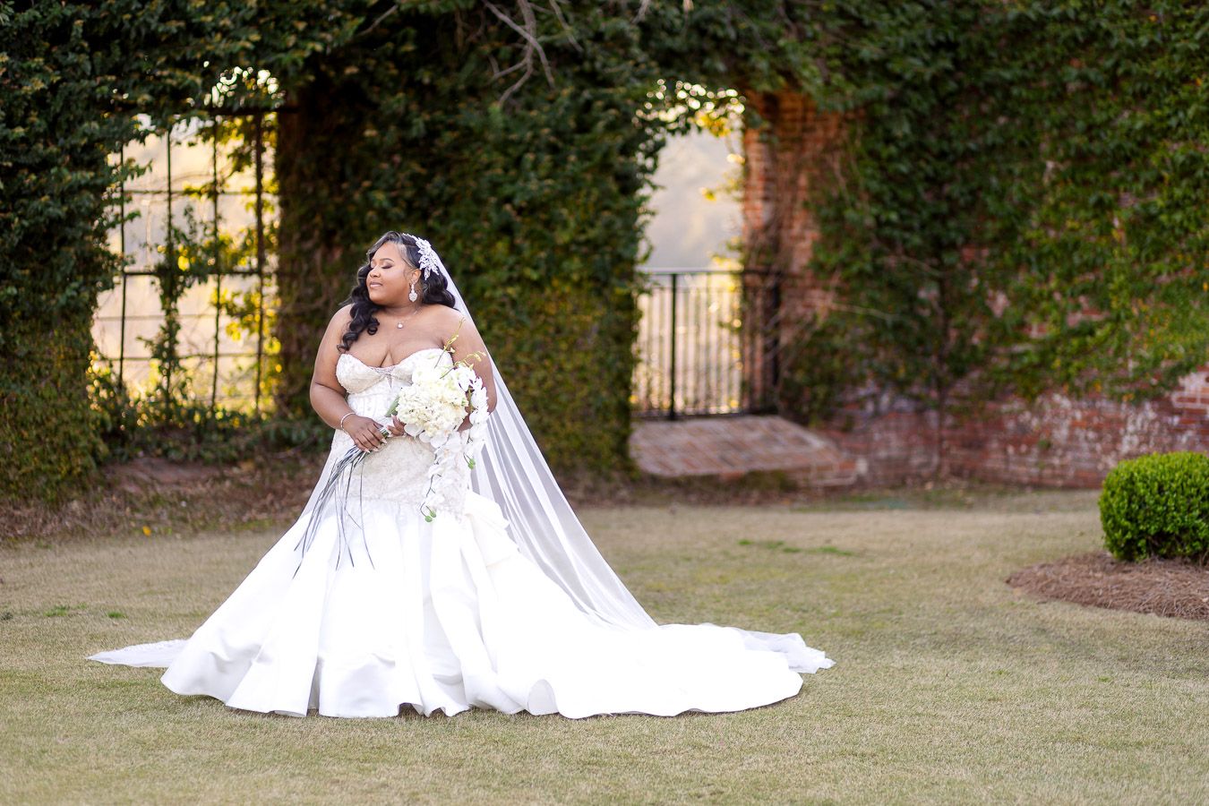 A person in an elegant white wedding gown stands gracefully on a manicured lawn with a stone archway in the background.
