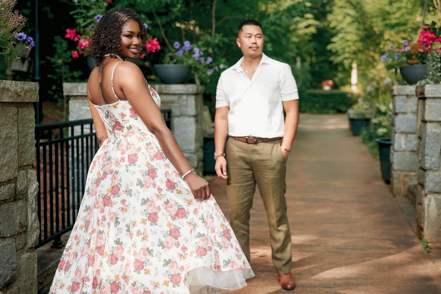 A couple poses on a stone pathway with hanging flower baskets, the person in a floral dress looking back over their shoulder.