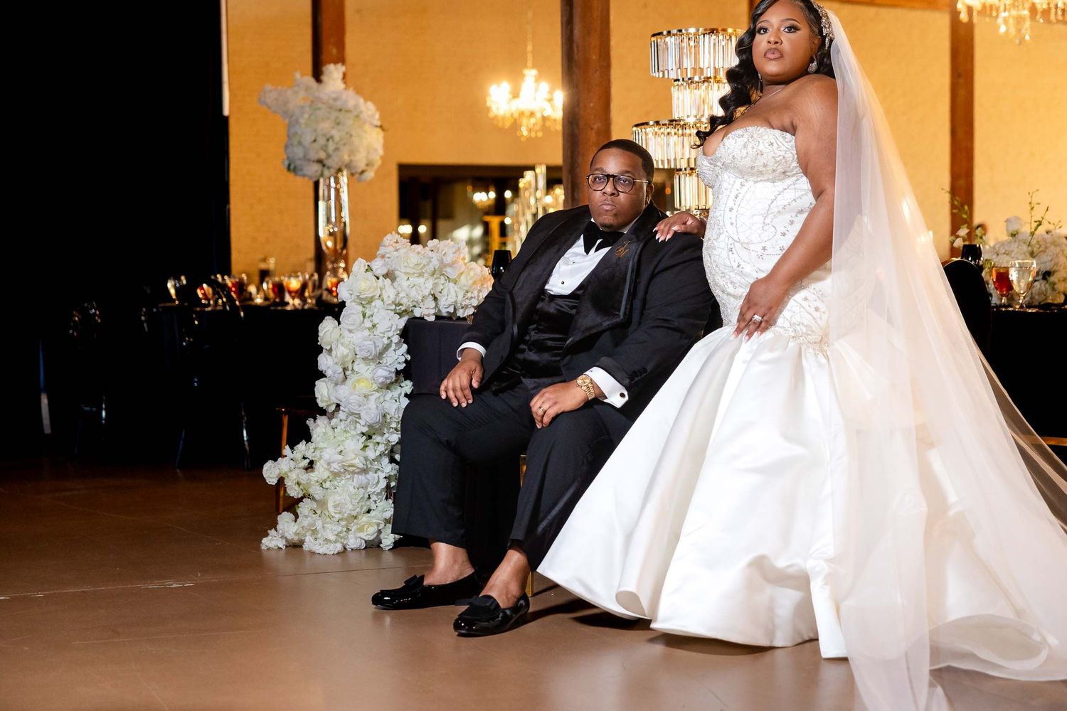 Elegant wedding scene with bride in white gown and groom in suit seated beside floral decorations in grand ballroom.