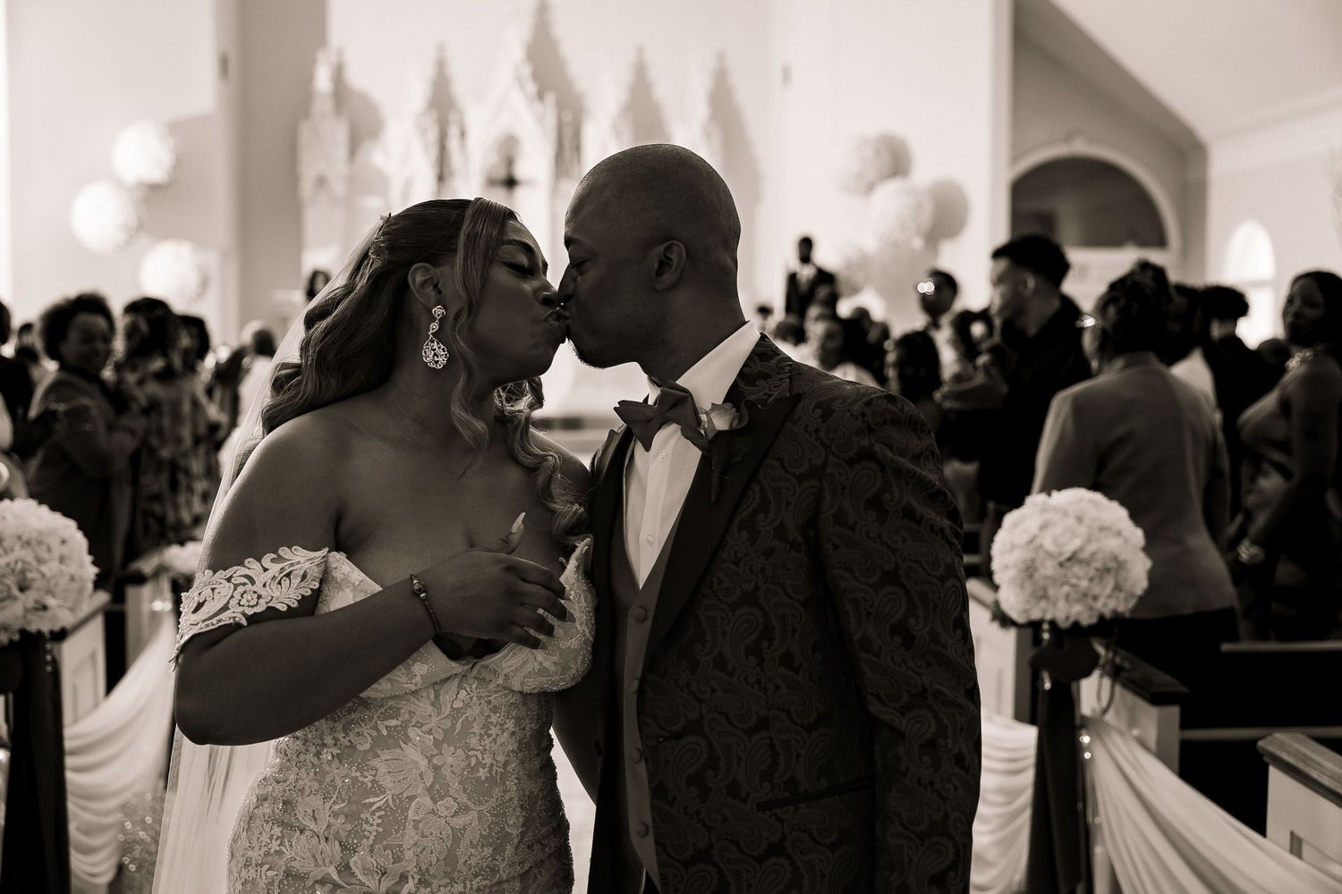 A romantic wedding moment captured in black and white inside a chapel adorned with white floral arrangements.