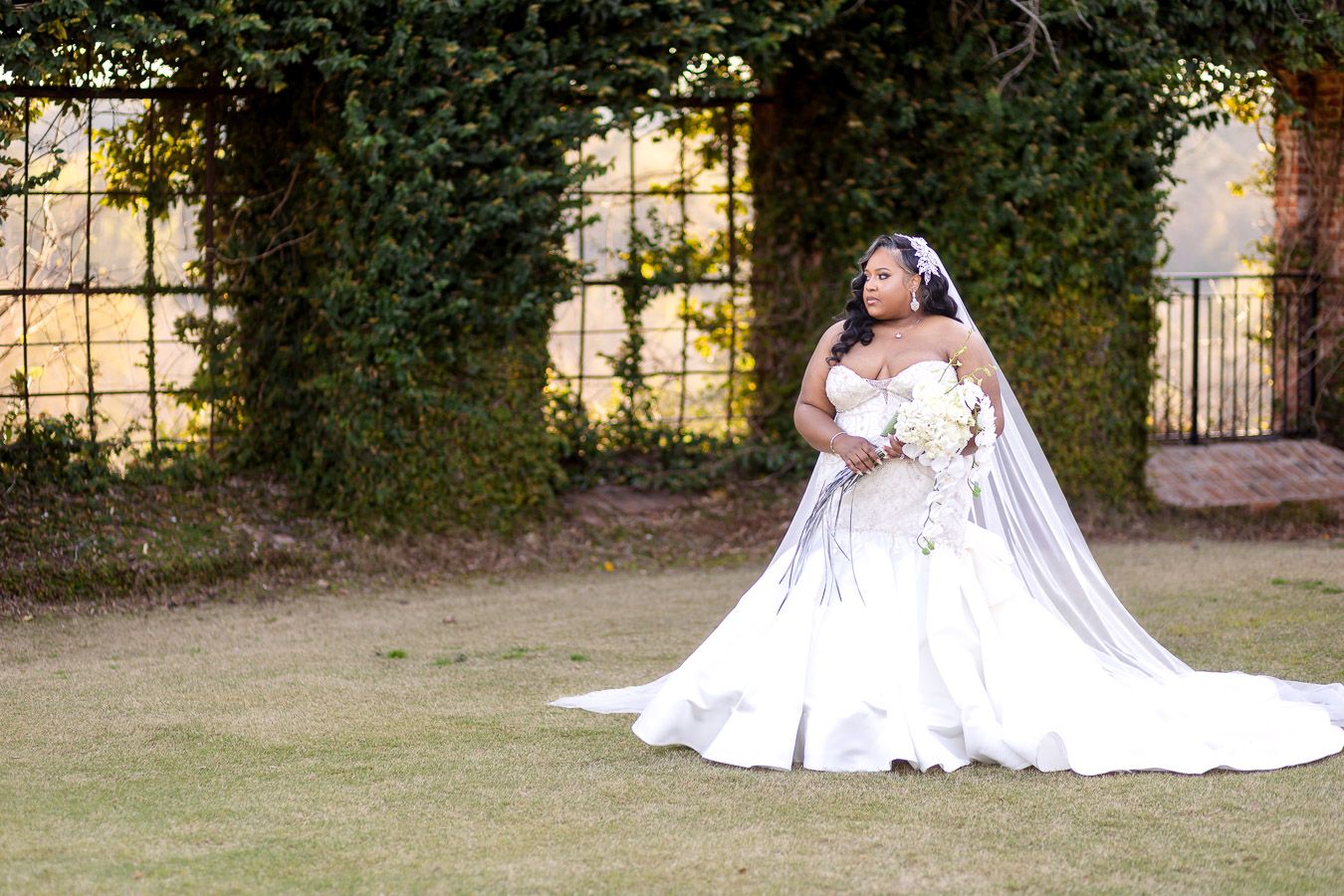 A person in an elegant white wedding dress and long veil sits gracefully on grass near an ivy-covered wall at golden hour.