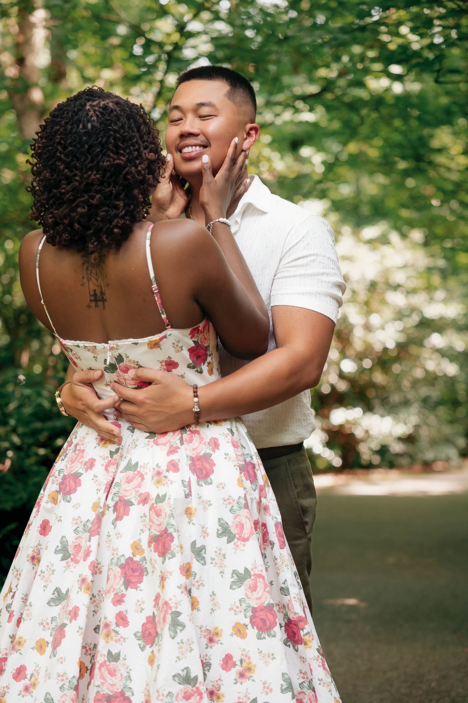 A romantic couple embraces outdoors while wearing a floral dress and white shirt against a lush green background.