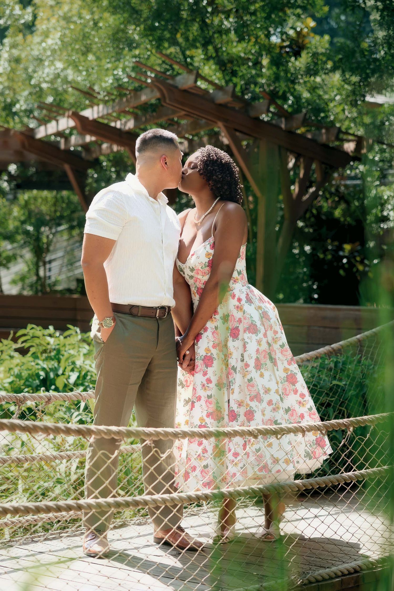 Couple in romantic attire share tender moments while walking along a rustic garden fence in dappled sunlight.