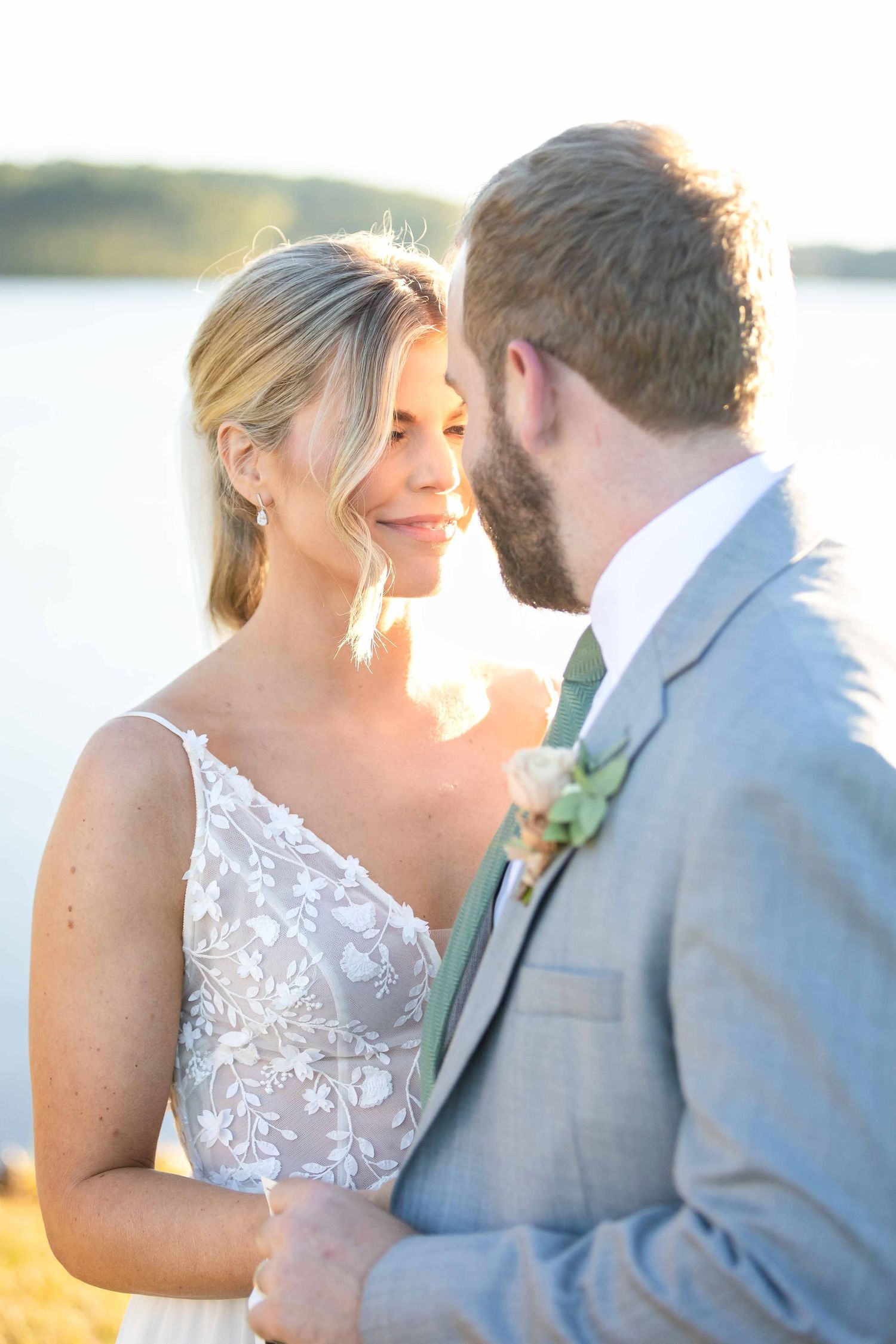 A romantic waterfront wedding photo of couple in white dress and grey suit during golden hour sunset.