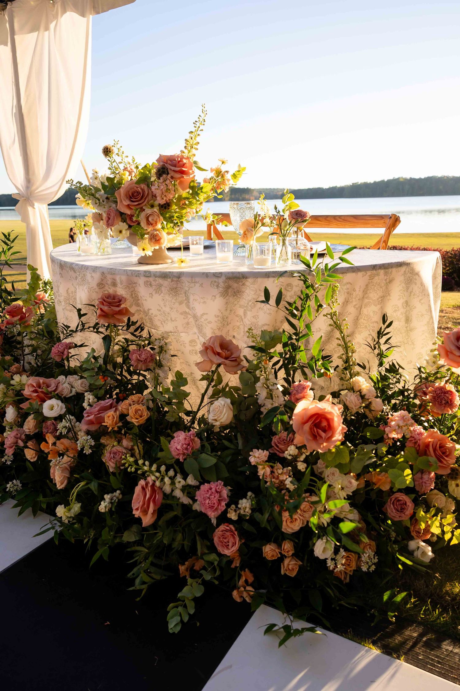 Elegant outdoor wedding table setting with lush floral arrangements in coral and pink tones against waterfront backdrop at sunset.