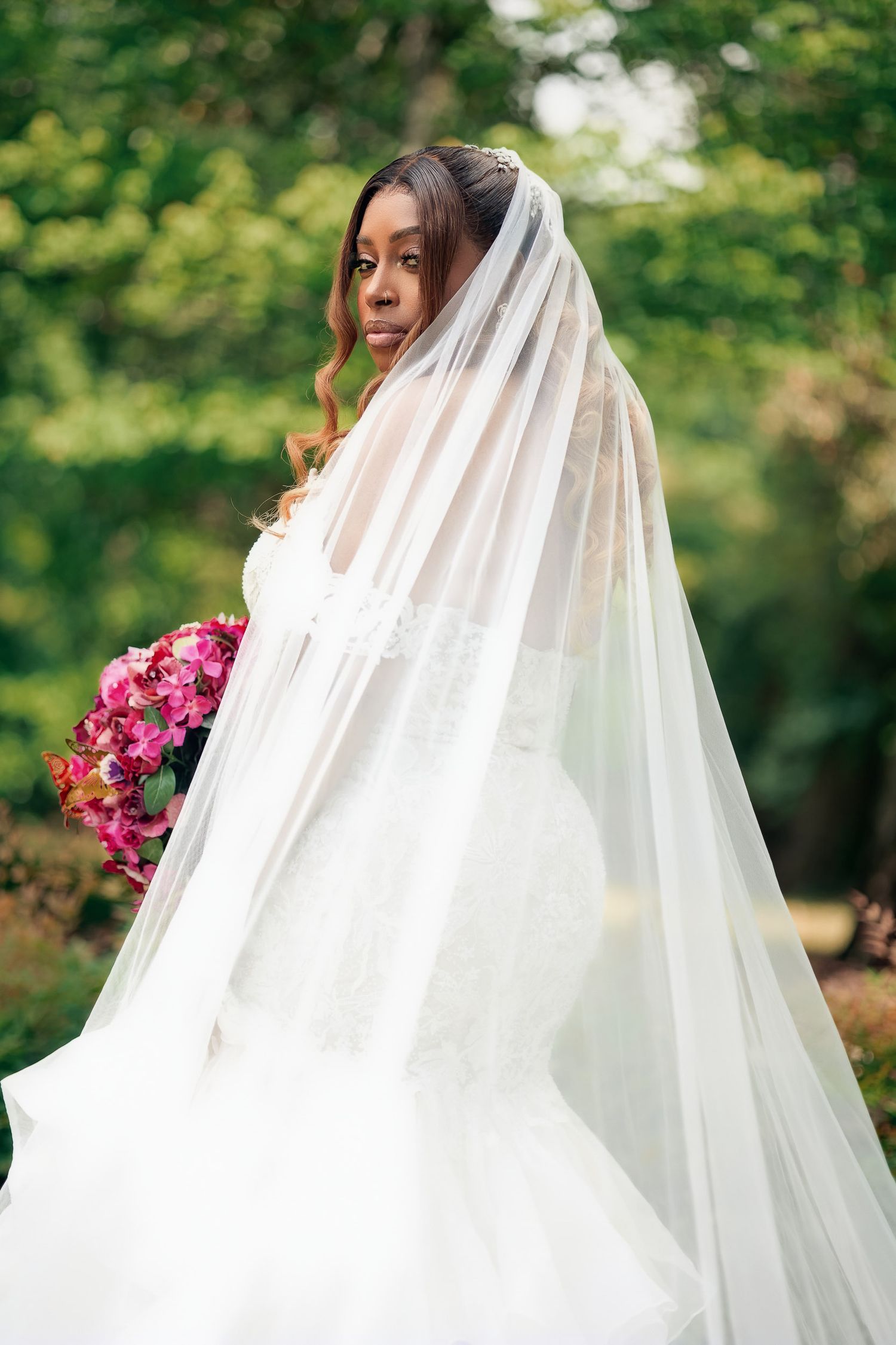 A bride in a flowing white wedding dress and veil holds a vibrant pink bouquet against a blurred green outdoor backdrop.