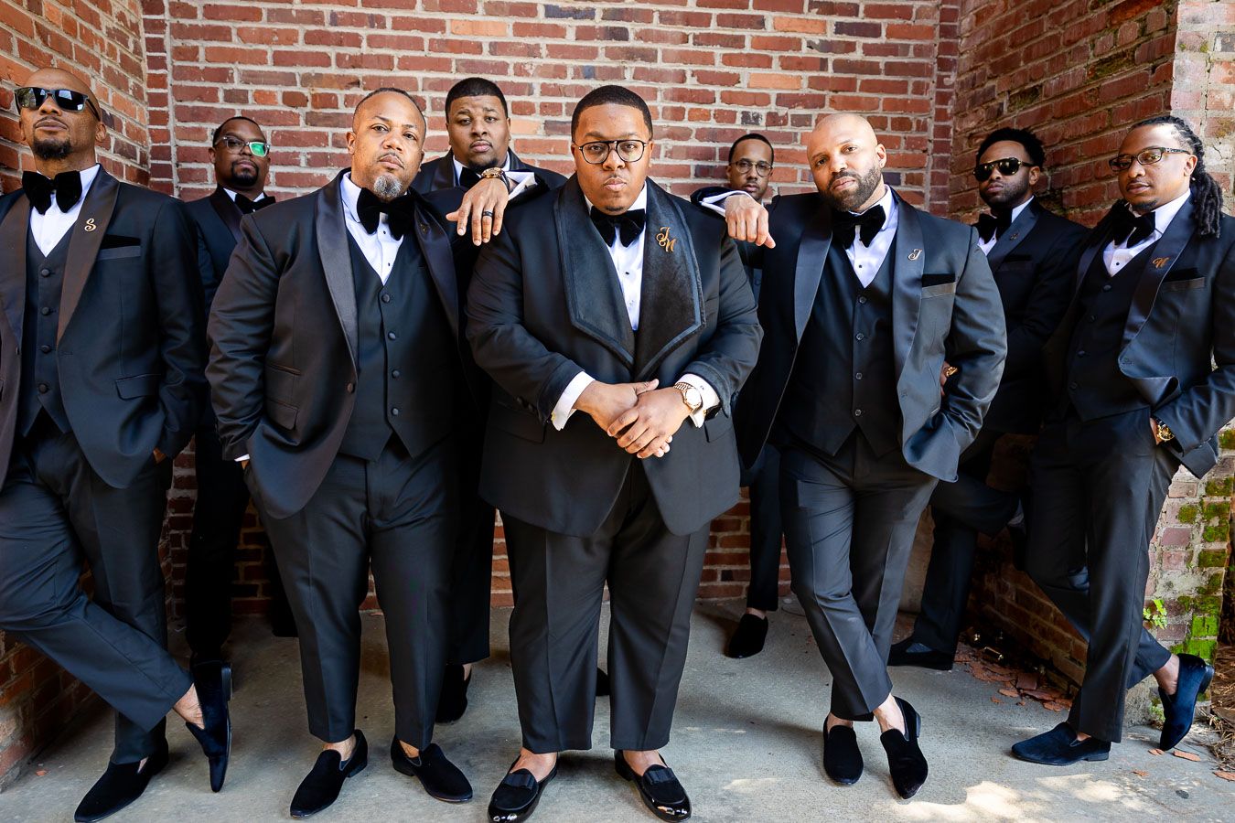 A group in formal black tuxedos and bow ties pose together against a brick wall for a classy portrait.