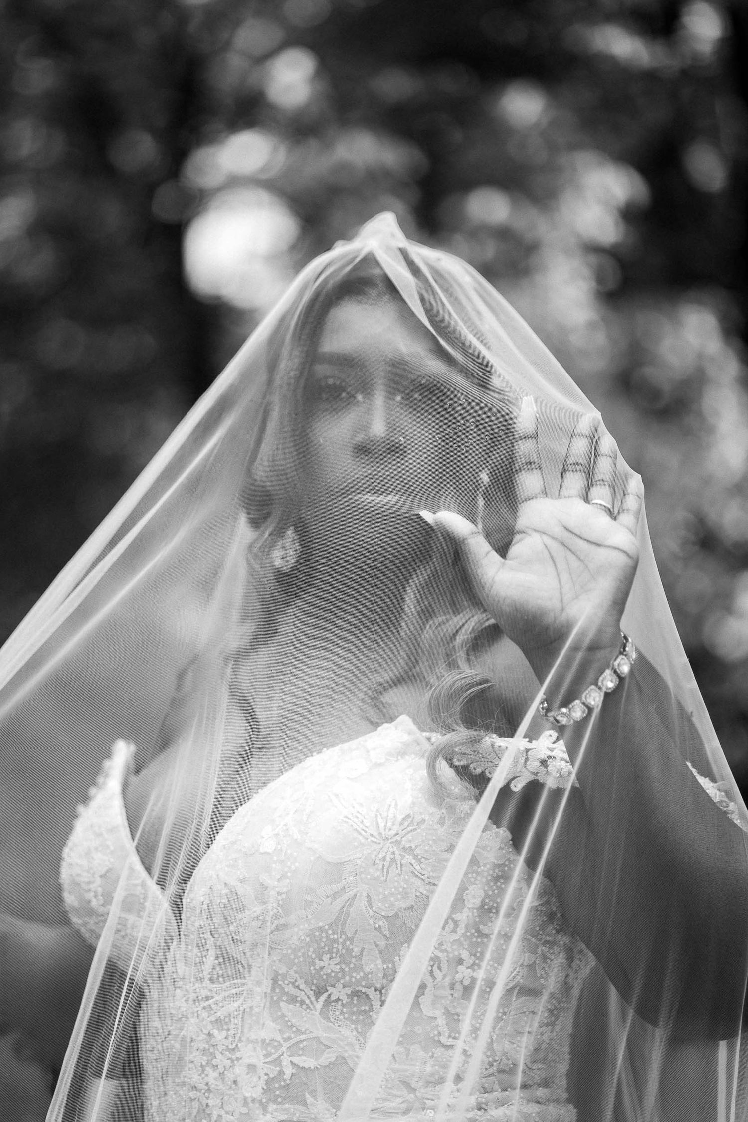 A bride in a white lace wedding dress and veil poses with her hand raised in an ethereal black and white portrait.