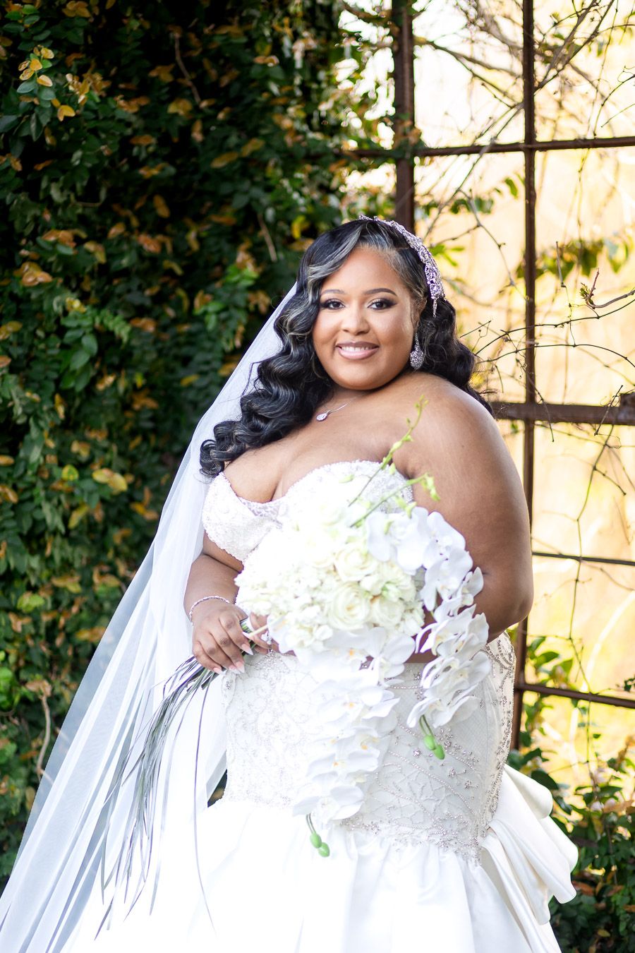 Elegant bride wearing white lace wedding gown with long veil and holding white floral bouquet in outdoor garden setting.