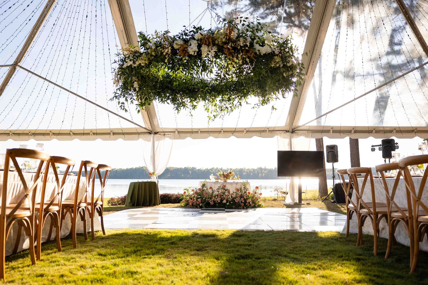 A wedding tent with wooden cross-back chairs and a floral chandelier overlooking the ocean on a sunny day.