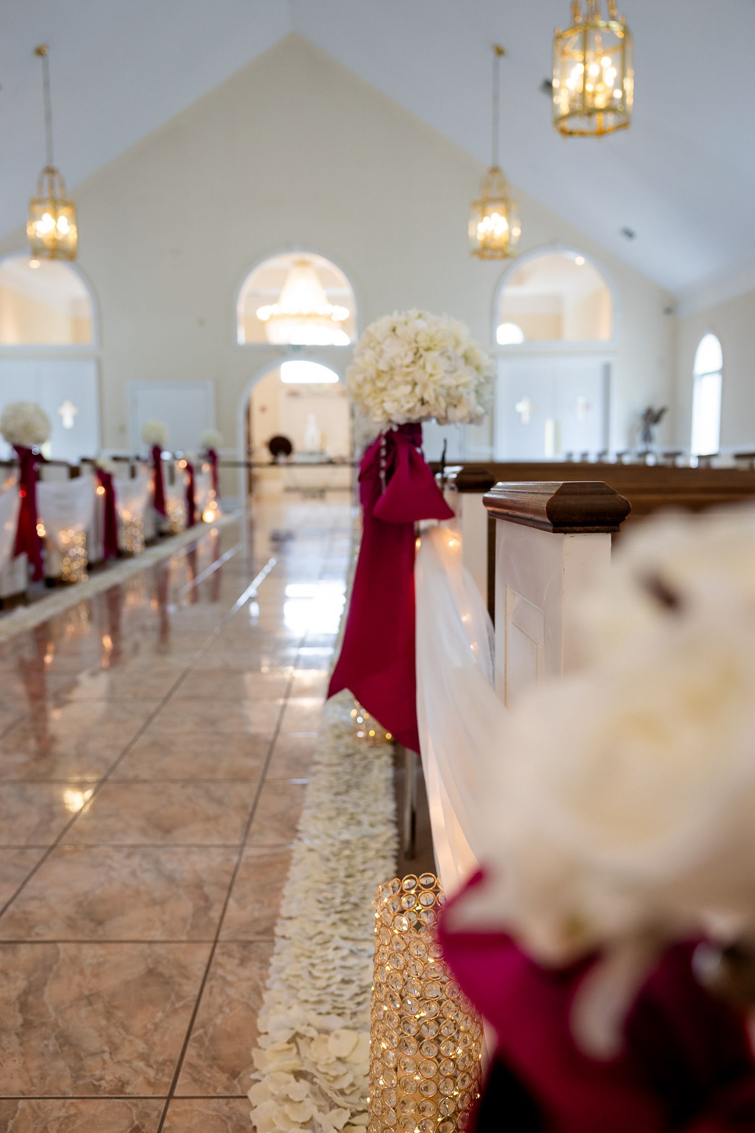 Elegant chapel interior with burgundy ribbon aisle decor, hanging lanterns, and white flower arrangements on pedestals.