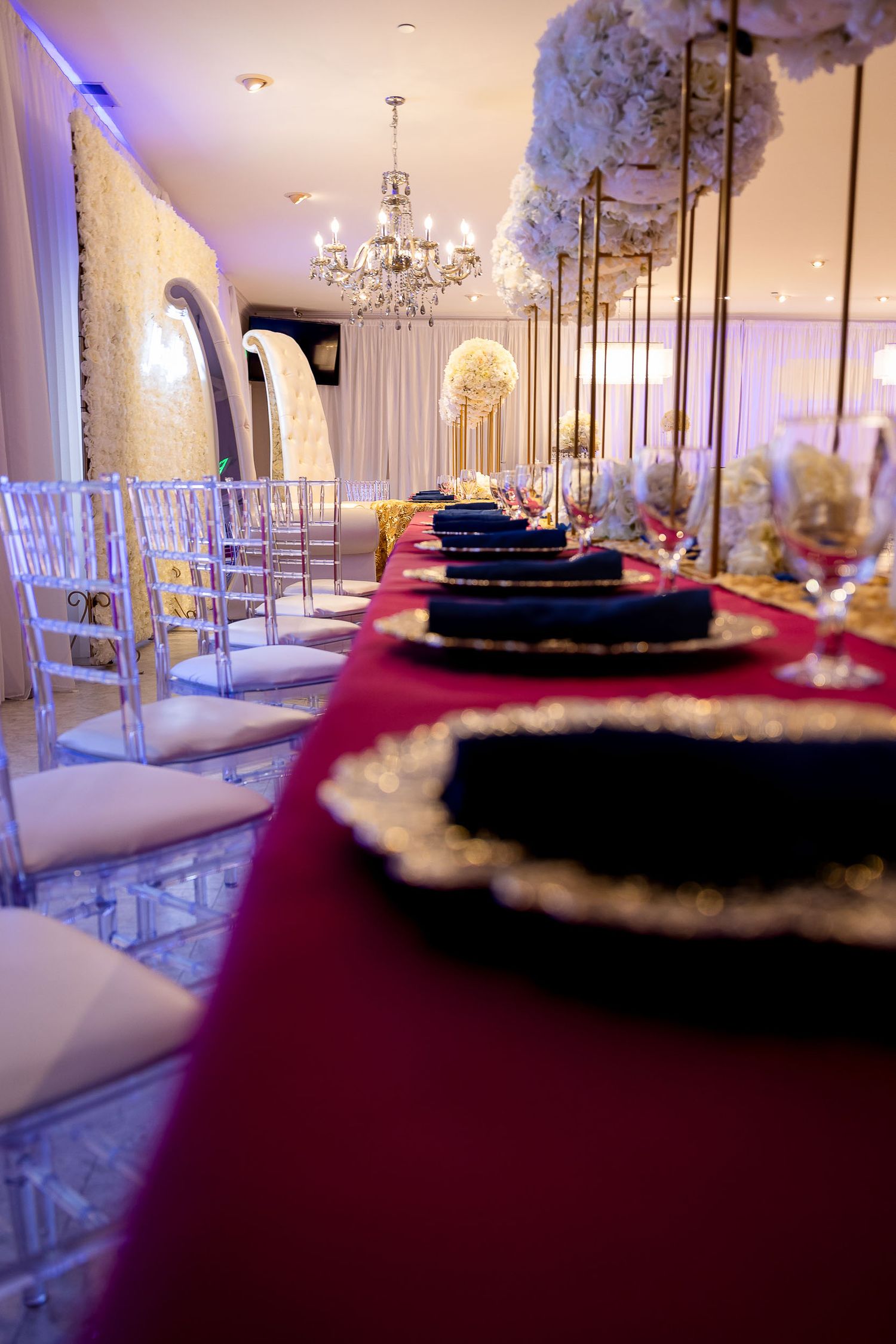 Elegant wedding reception table with red tablecloth, crystal chandelier, and clear chiavari chairs in a luxurious ballroom.