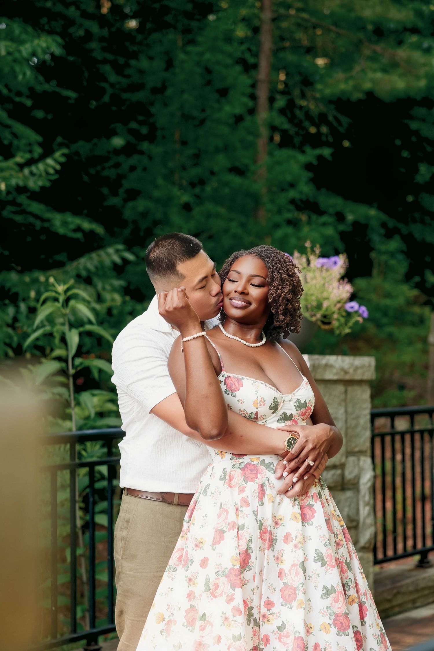 A couple shares romantic moments in an outdoor setting with greenery background while wearing white and floral attire.