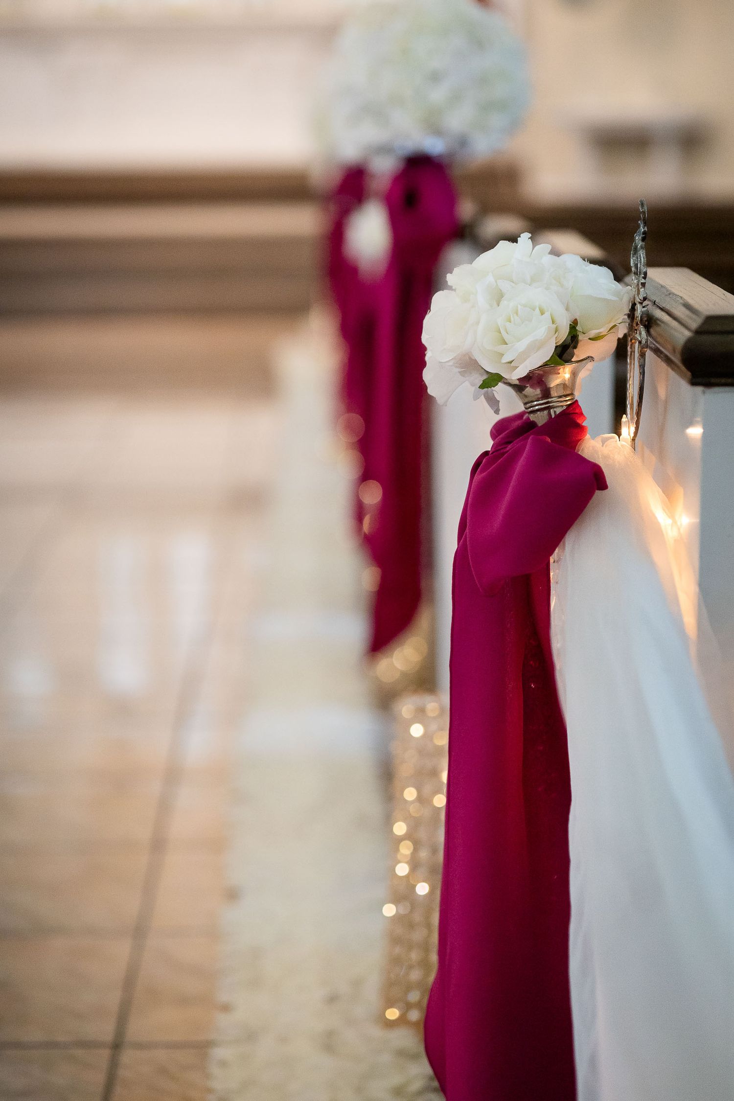 White rose floral arrangements with burgundy fabric draping and twinkling lights decorate an indoor wedding venue aisle.