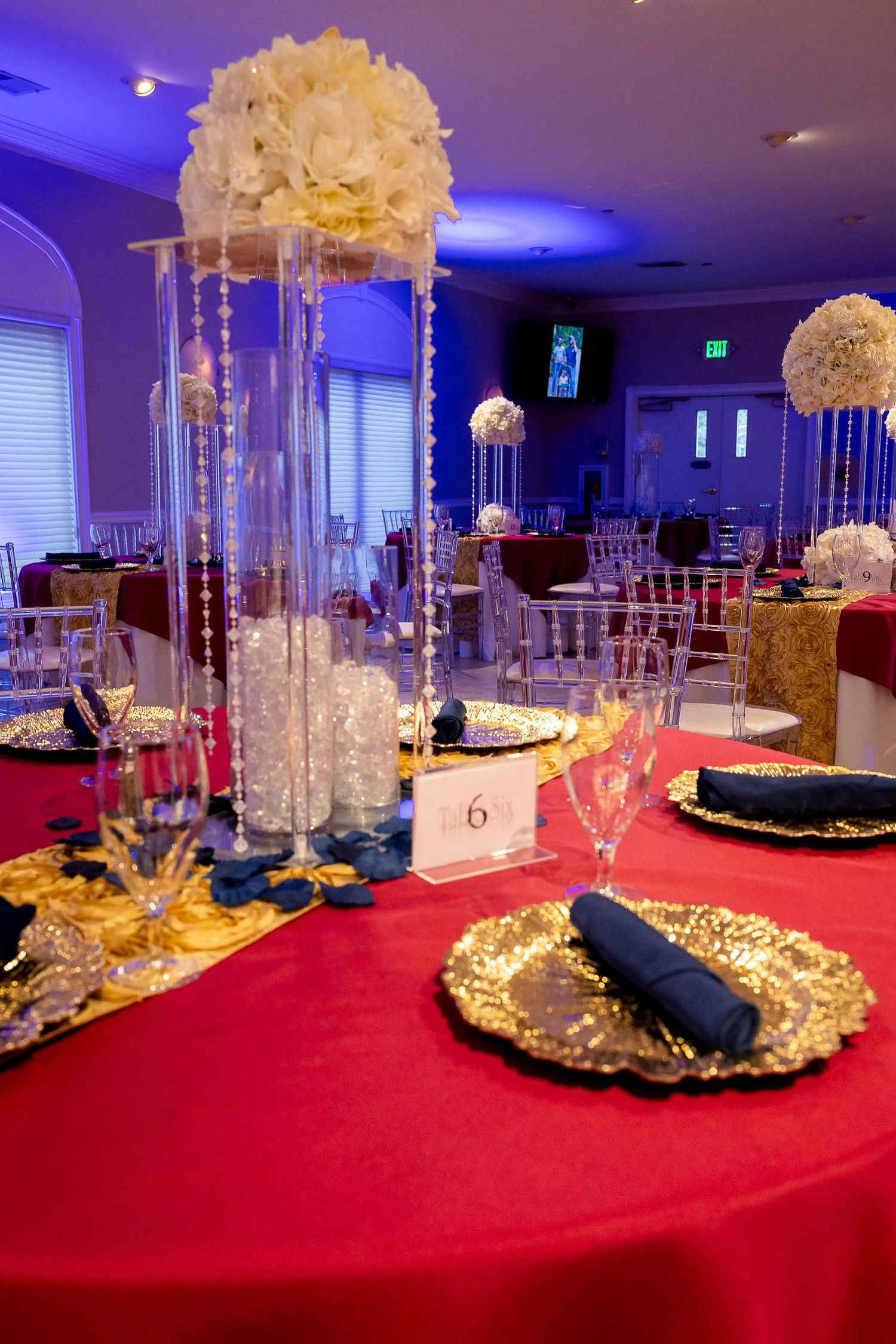 Elegant wedding reception table with red tablecloth, gold chargers, and tall white floral centerpieces under blue lighting.