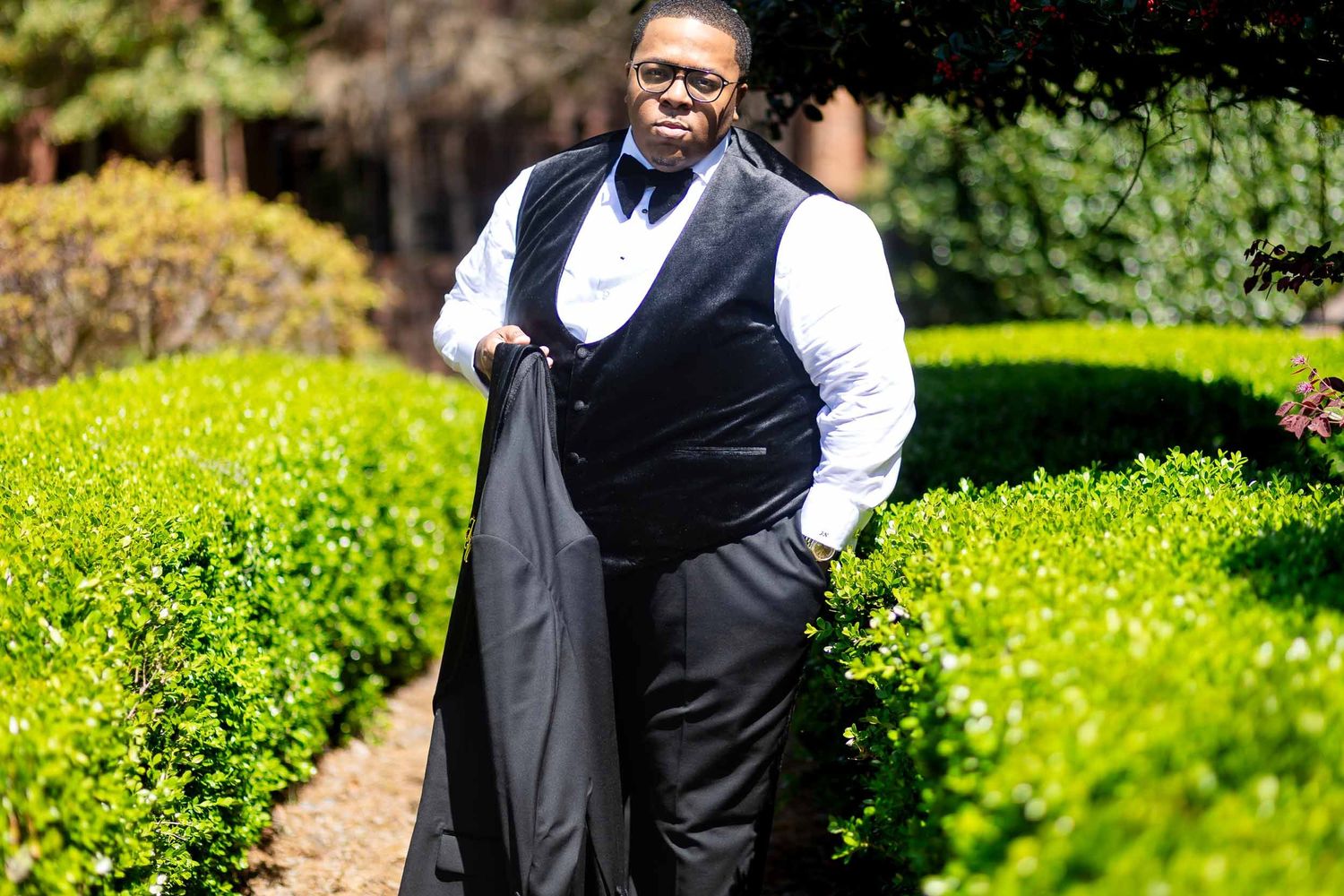 Someone wearing formal attire with black vest and white dress shirt smiles in a garden path with green hedges.