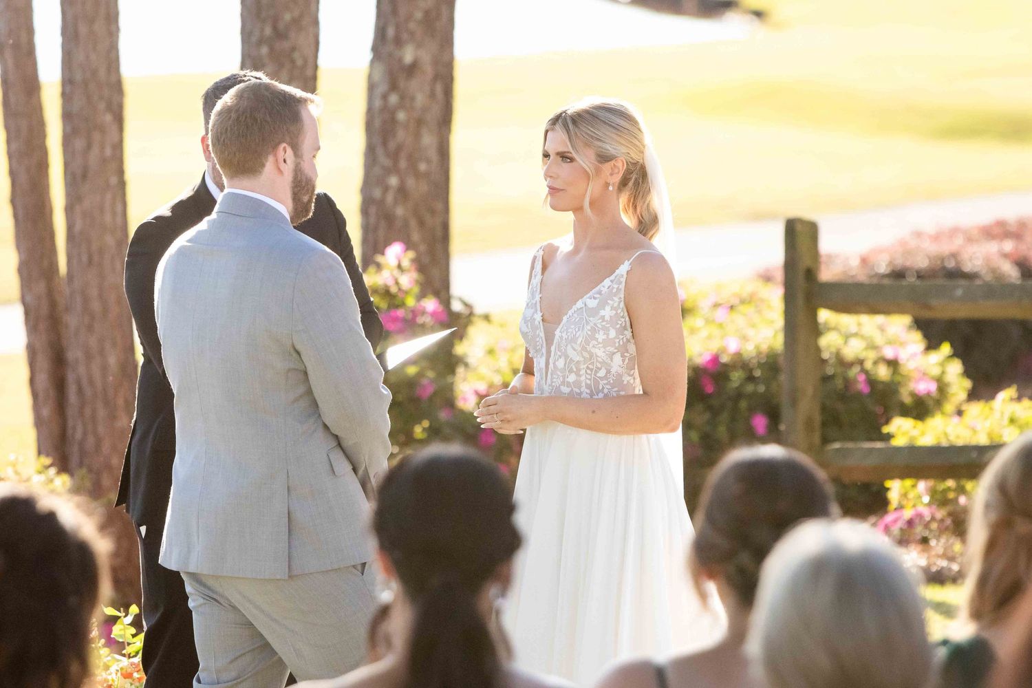 A romantic outdoor wedding ceremony takes place among tall pine trees during golden hour with guests watching.